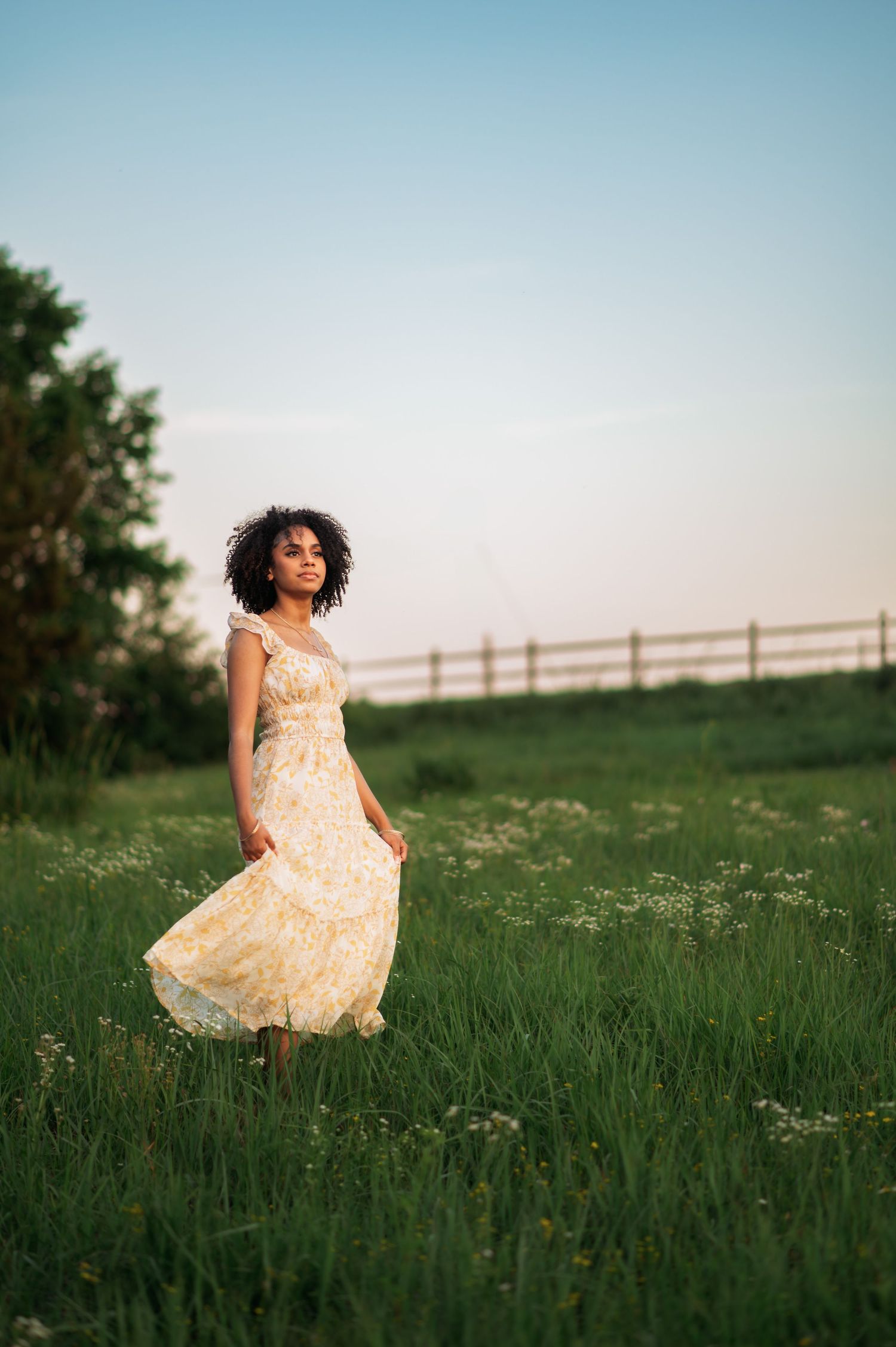 A person in a flowing yellow floral dress stands in a sunlit meadow with a wooden fence in the background at sunset.