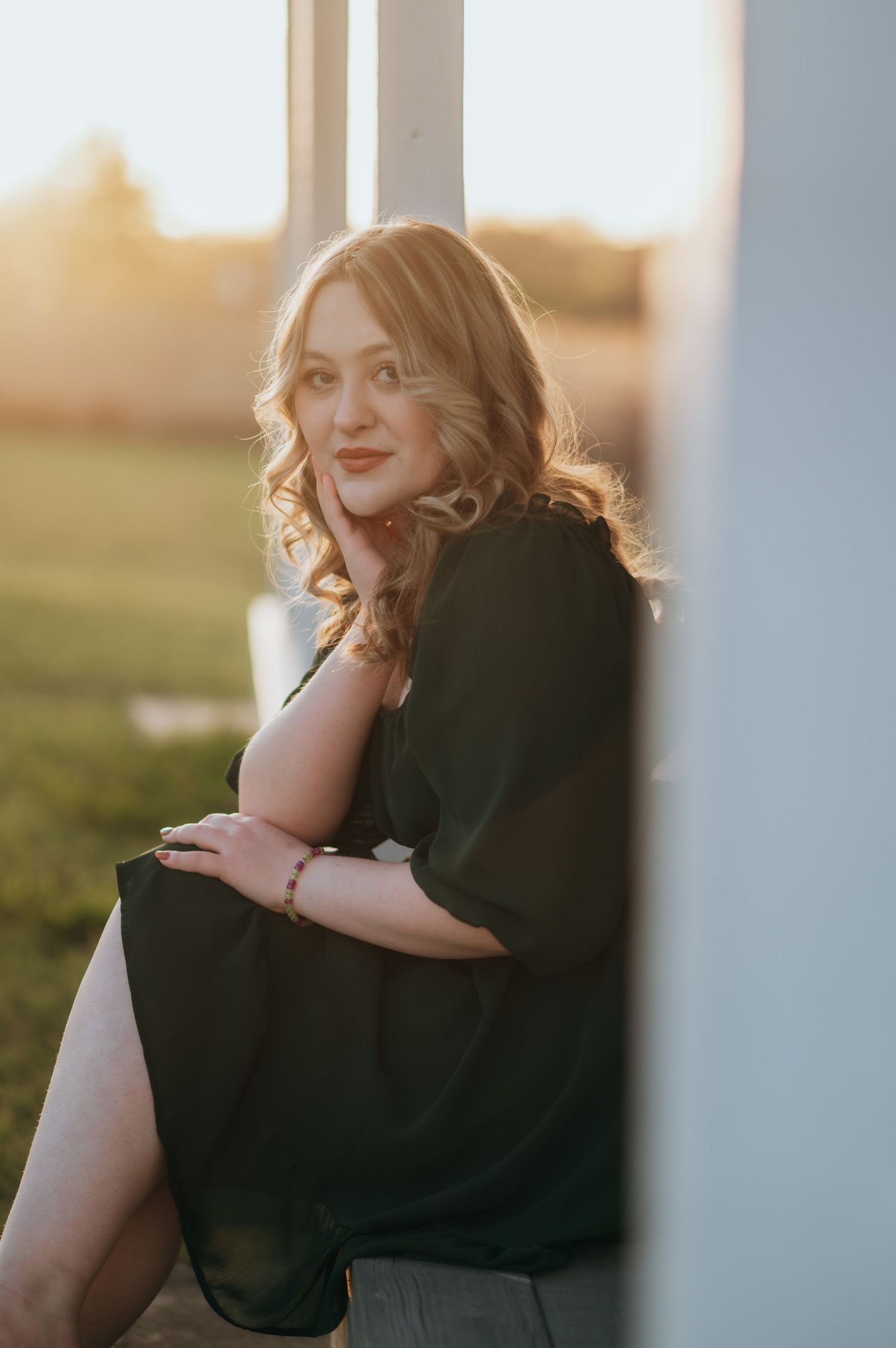 Woman in black dress sitting against white pillar at golden hour sunset, with curly blonde hair and warm glowing light.