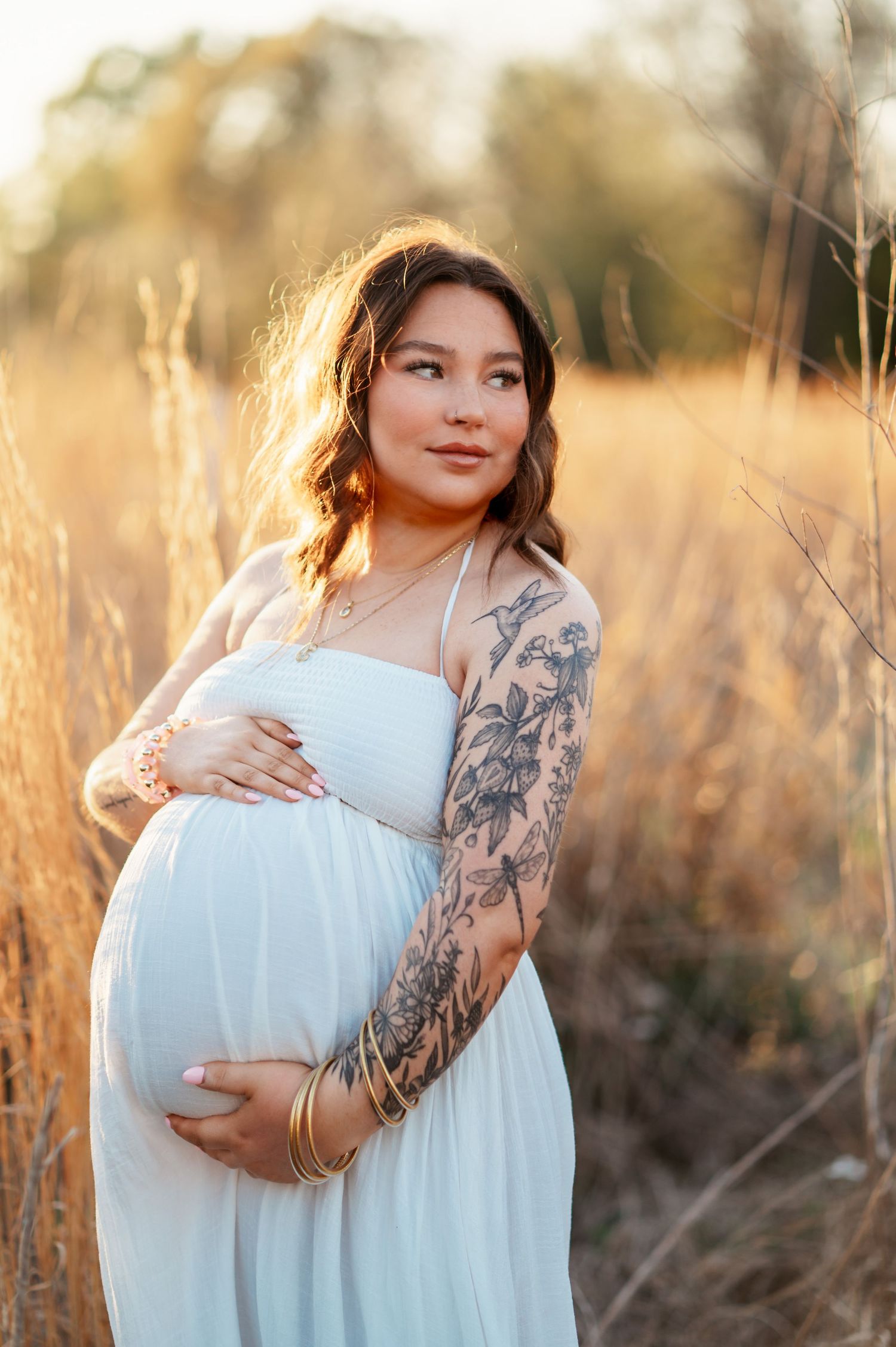 Pregnant woman in white dress with floral arm tattoo cradling baby bump in golden field at sunset.