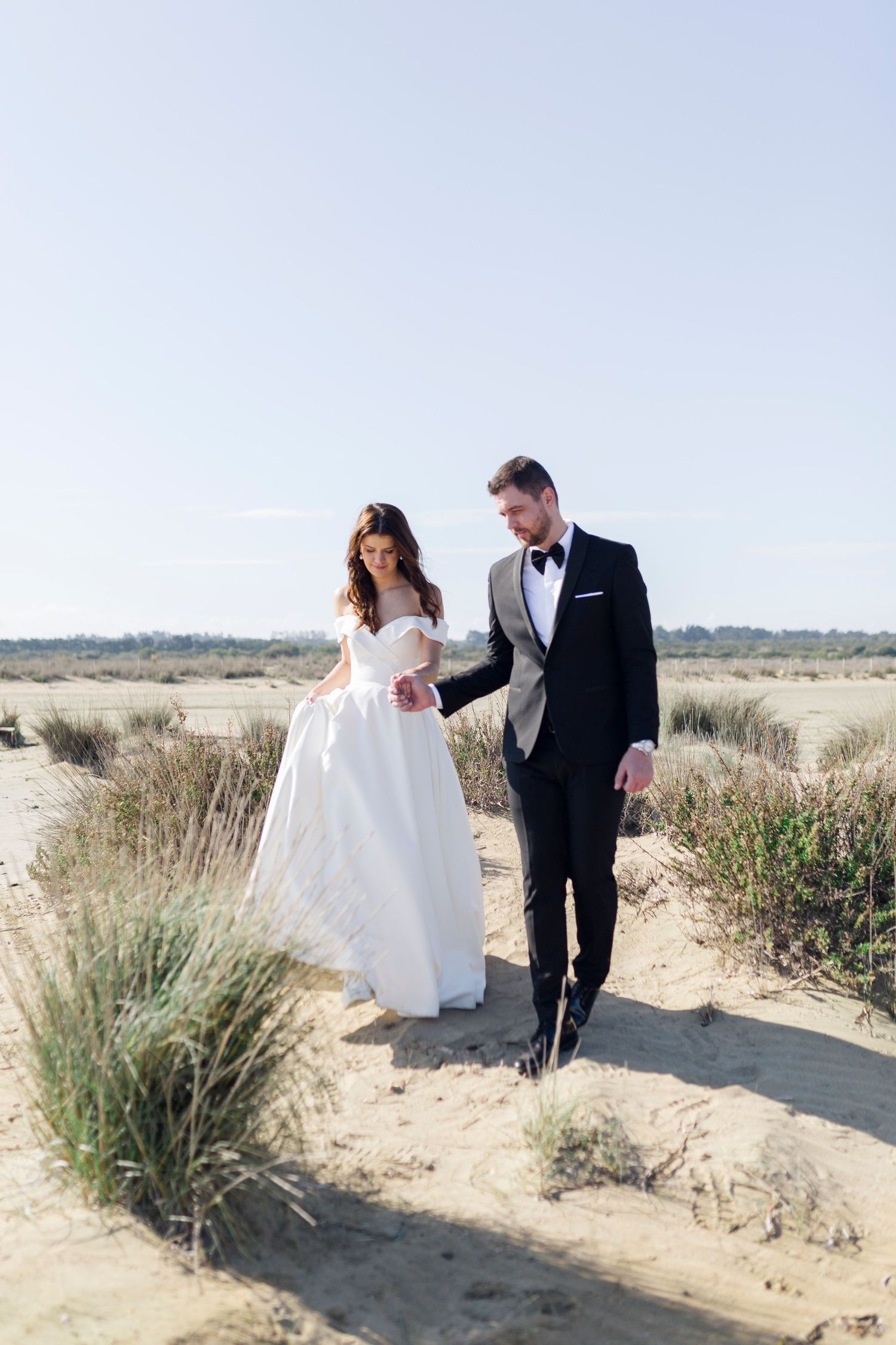 Bride and groom walking hand in hand on sandy dunes, bride in white gown, groom in black tuxedo with bow tie.