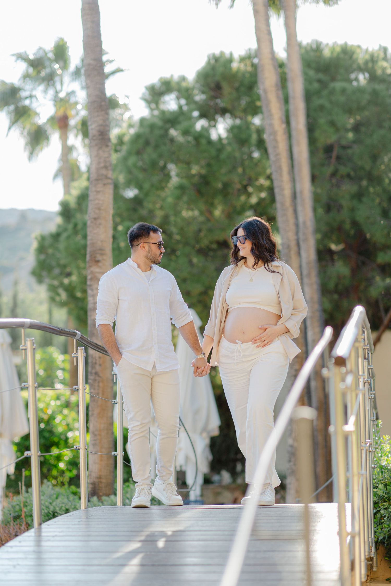 Pregnant couple holding hands walking on a bridge surrounded by palm trees, dressed in matching white outfits.