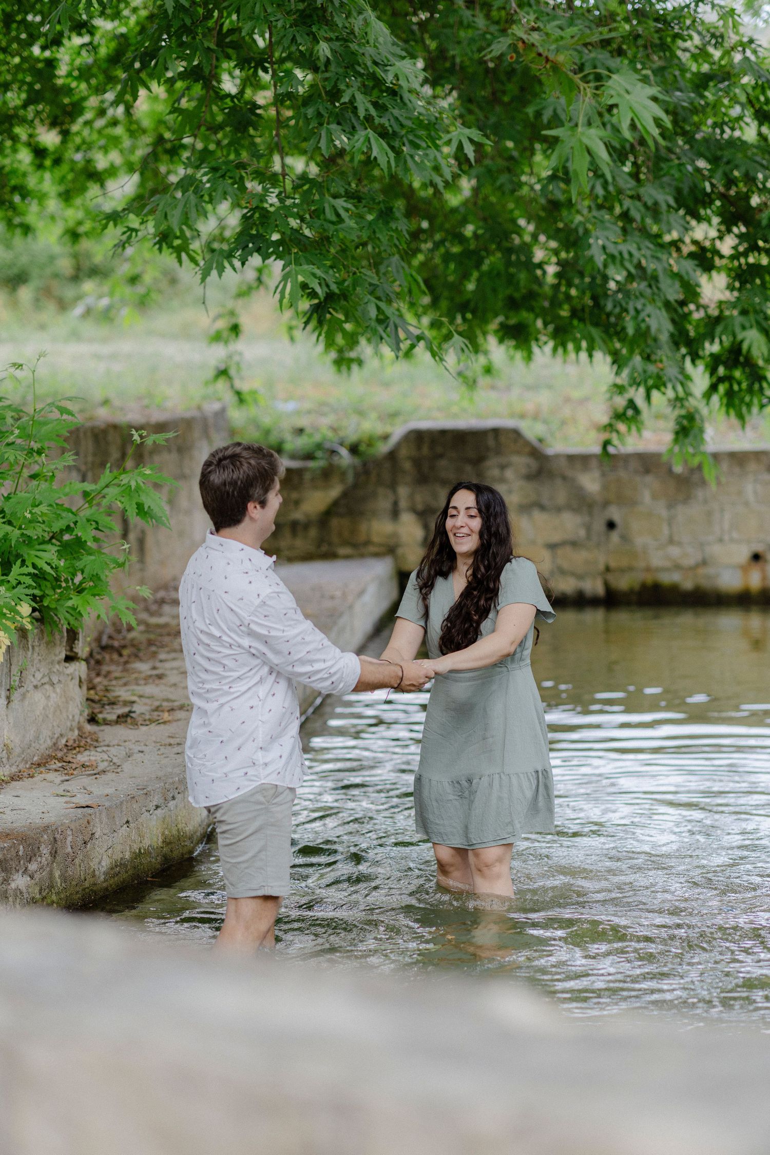 Couple holding hands and smiling while standing in a shallow creek, surrounded by lush green trees.