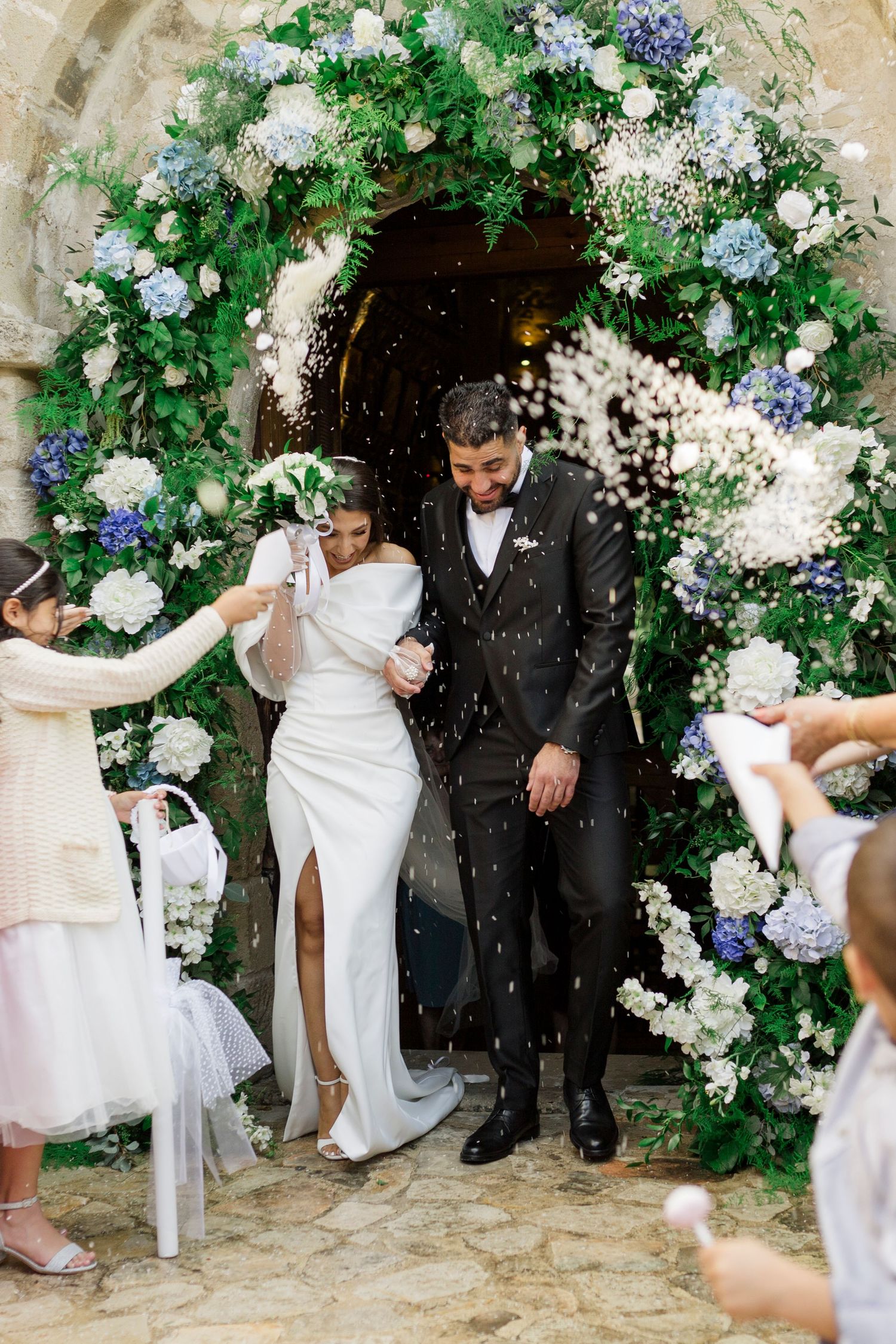 Newlyweds exit church ceremony under floral arch of blue hydrangeas as guests toss white confetti.
