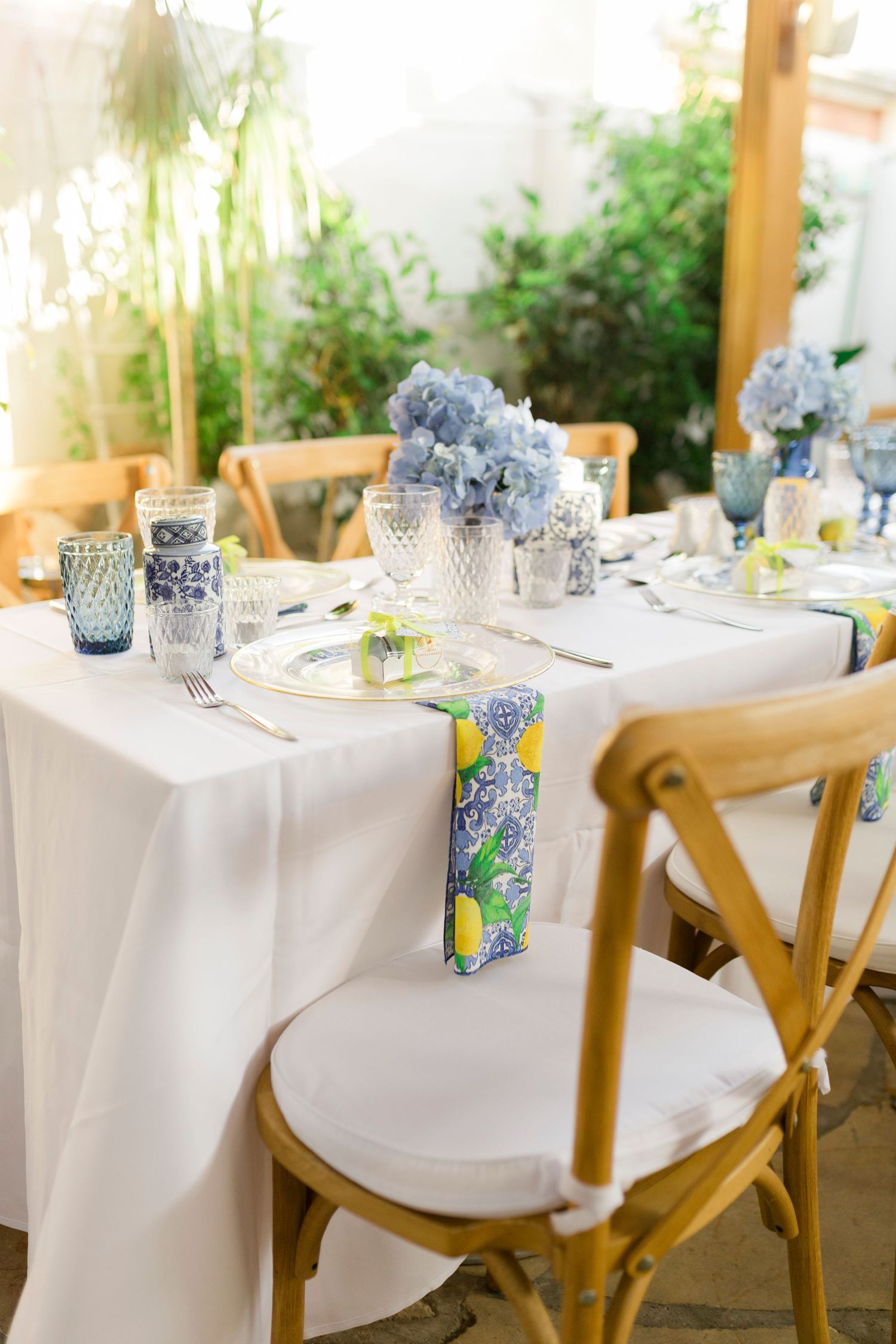 Elegant wedding table with white linen, blue hydrangeas, floral napkins, and wooden cross-back chairs in a garden setting.