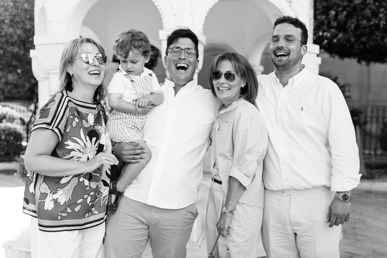 Black and white photo of a joyful family group of six laughing together outdoors near white arched architecture.