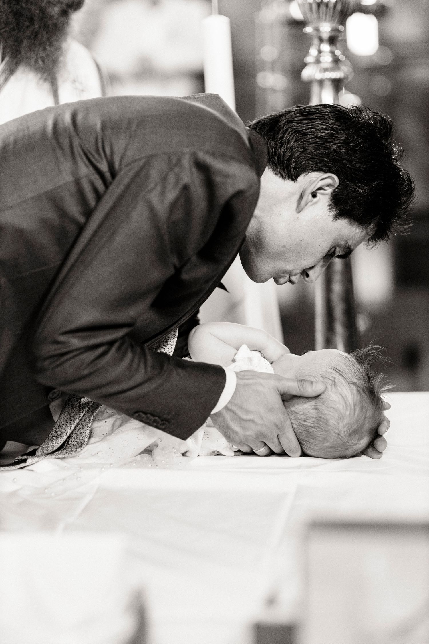 Man in suit tenderly kisses newborn baby lying on white surface during a christening ceremony, black and white photo.