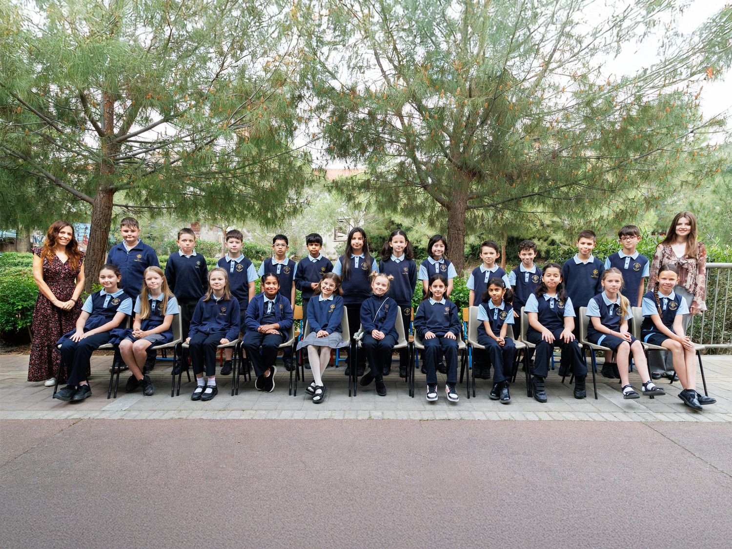 Group photo of school children in navy uniforms seated outdoors with teachers, trees in background.