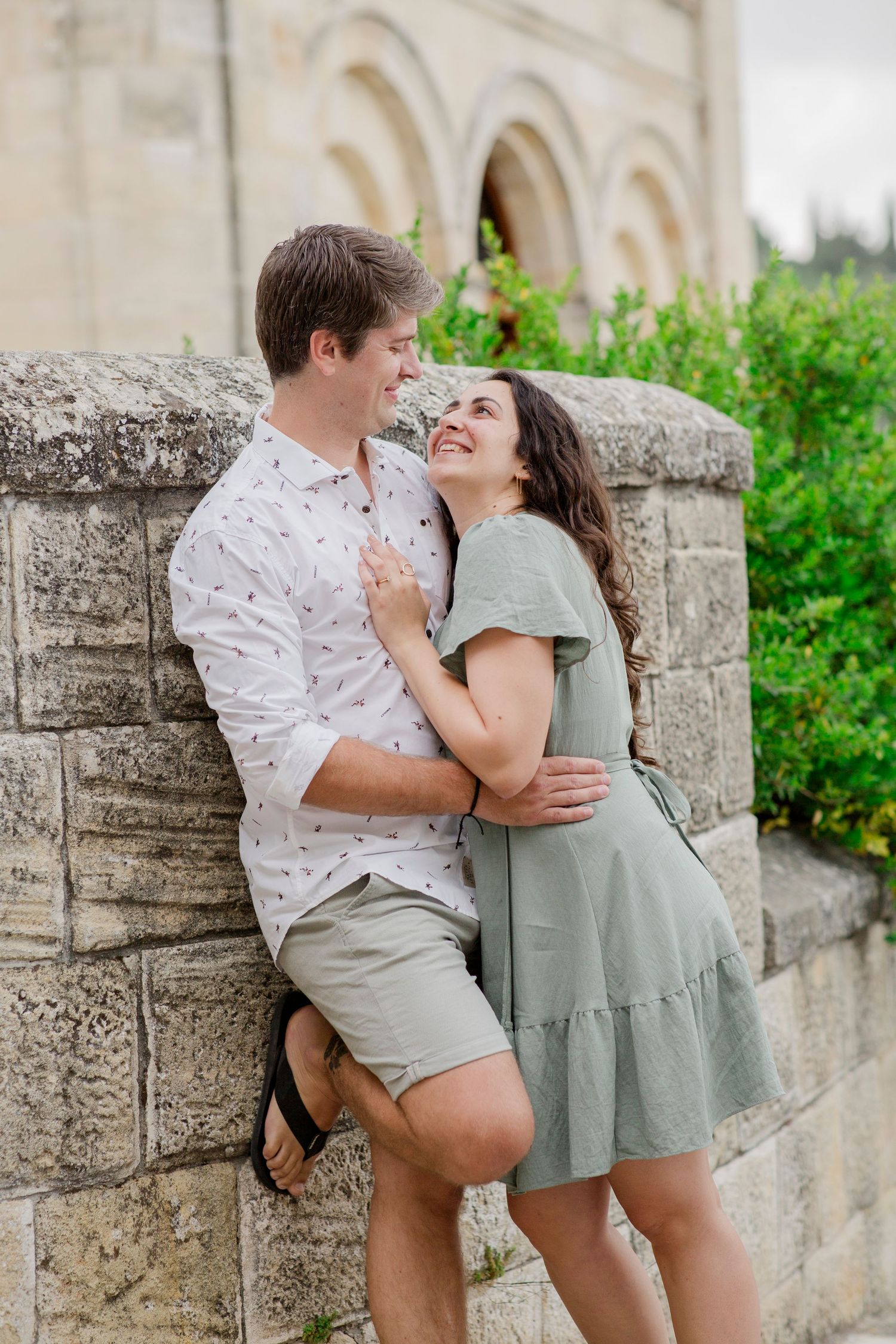 A smiling couple embraces against a stone wall, the woman in a sage green dress and man in a white shirt and khaki shorts.