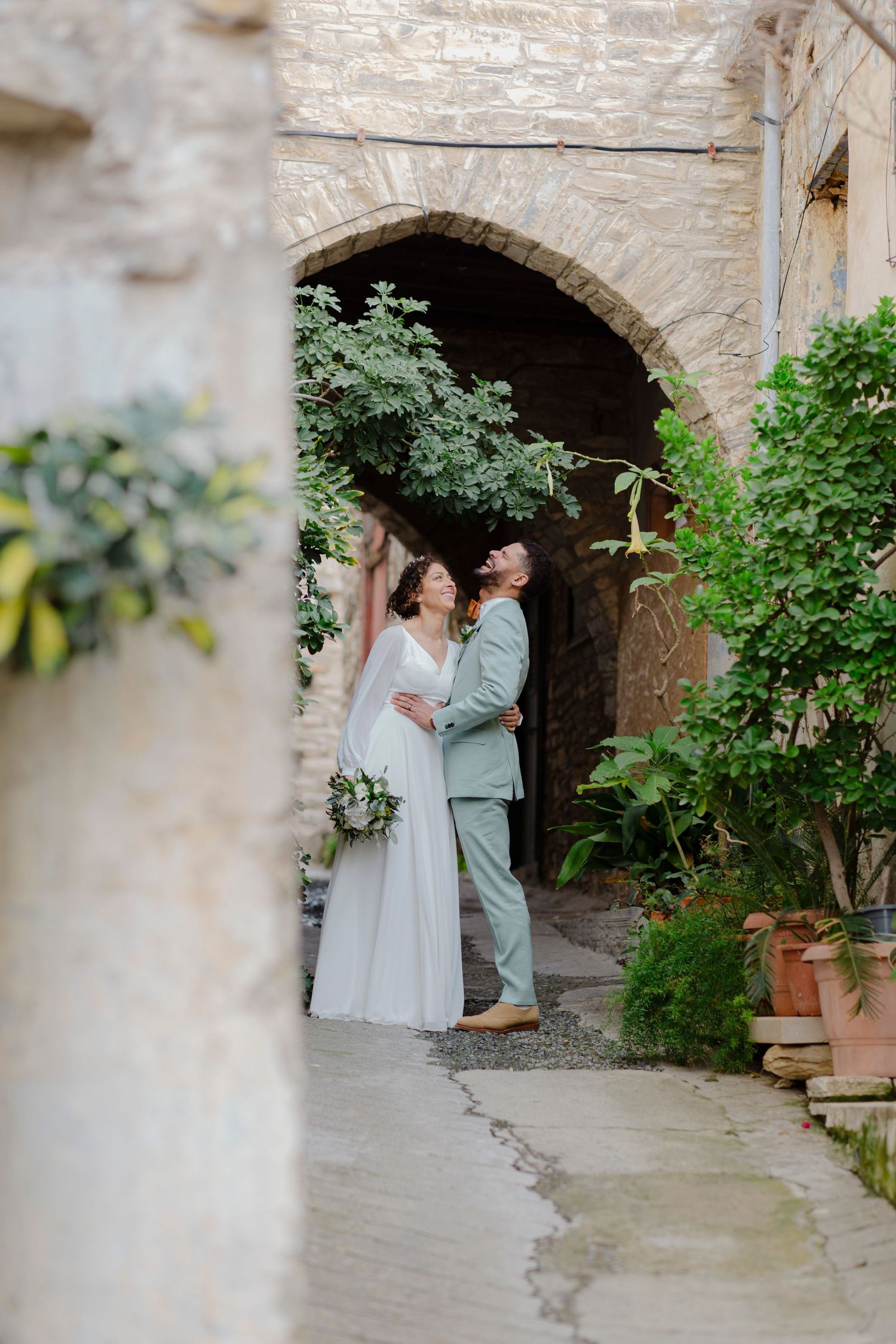 Bride and groom embrace under stone arch surrounded by lush greenery at rustic wedding venue.