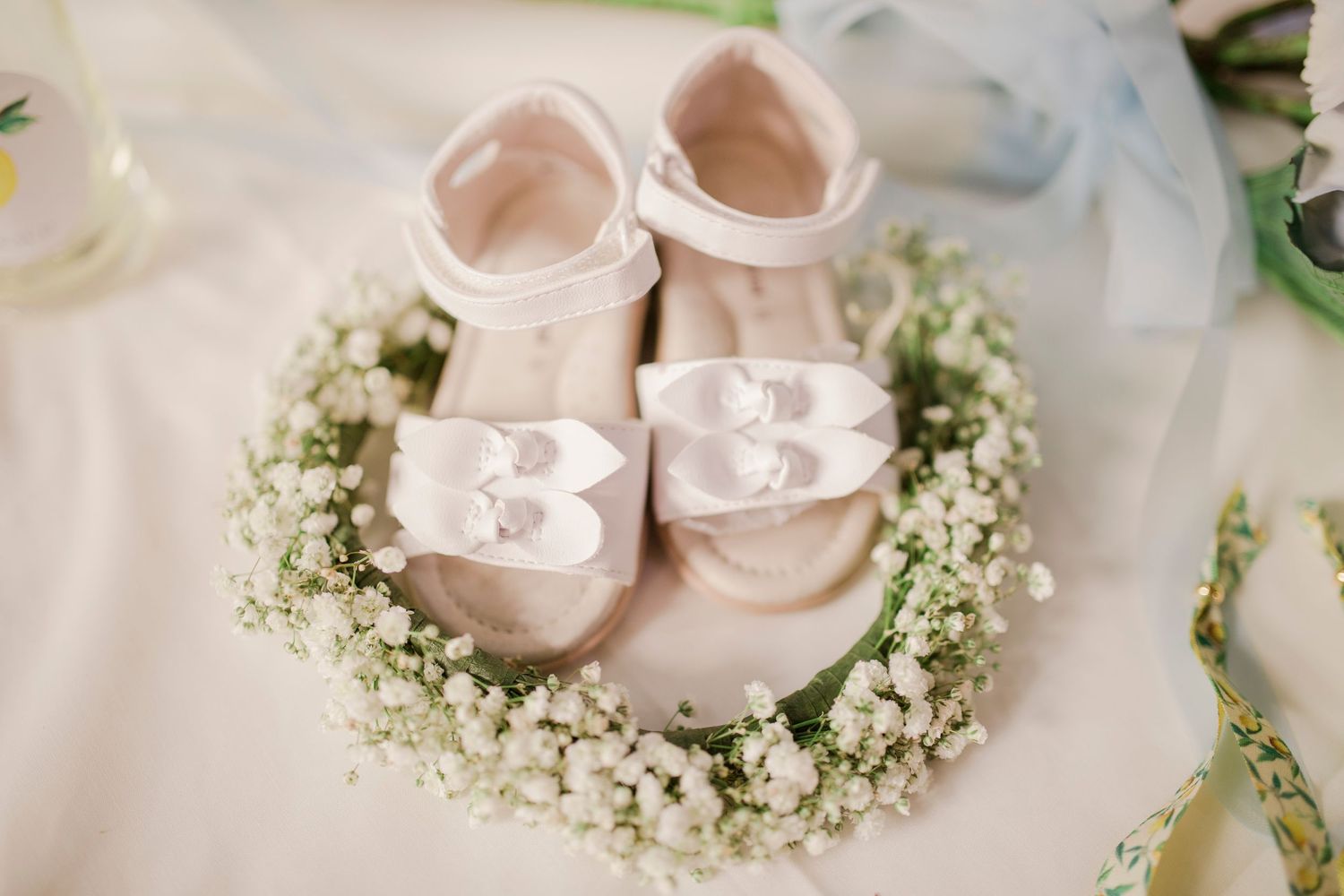 White baby shoes with bow details resting in a delicate wreath of baby's breath flowers on a white fabric.