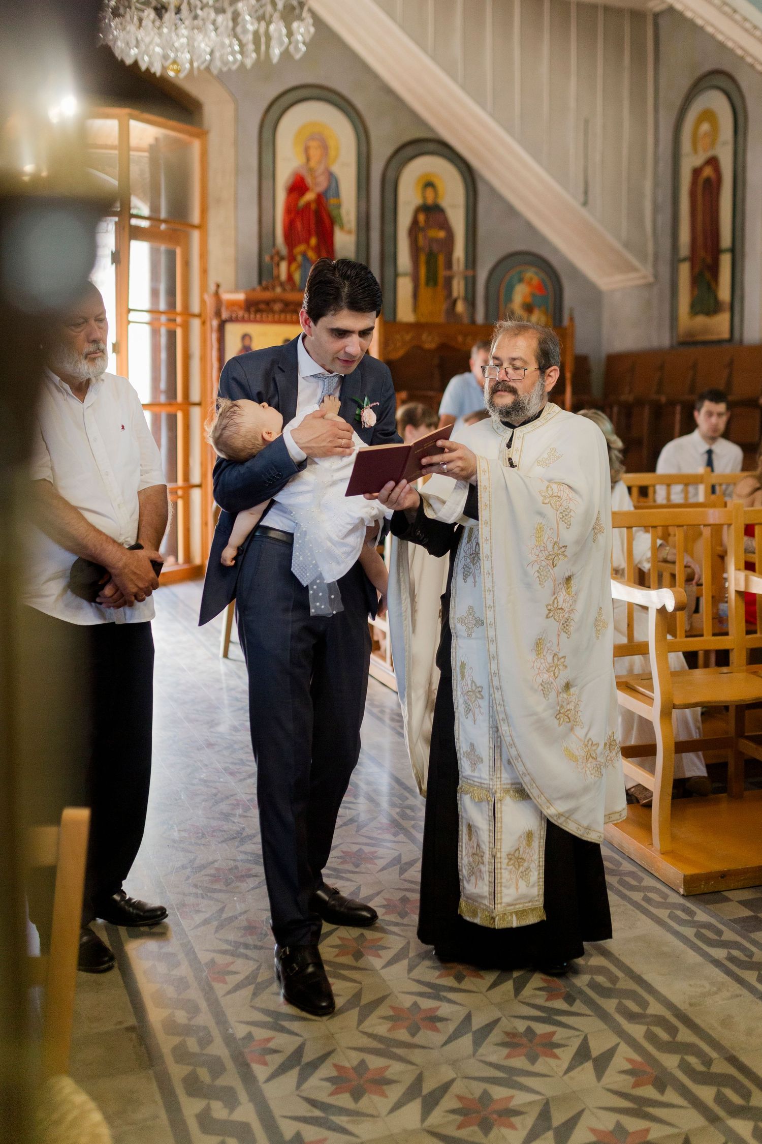 Priest and godfather holding infant during Orthodox Christian baptism ceremony inside decorated church.