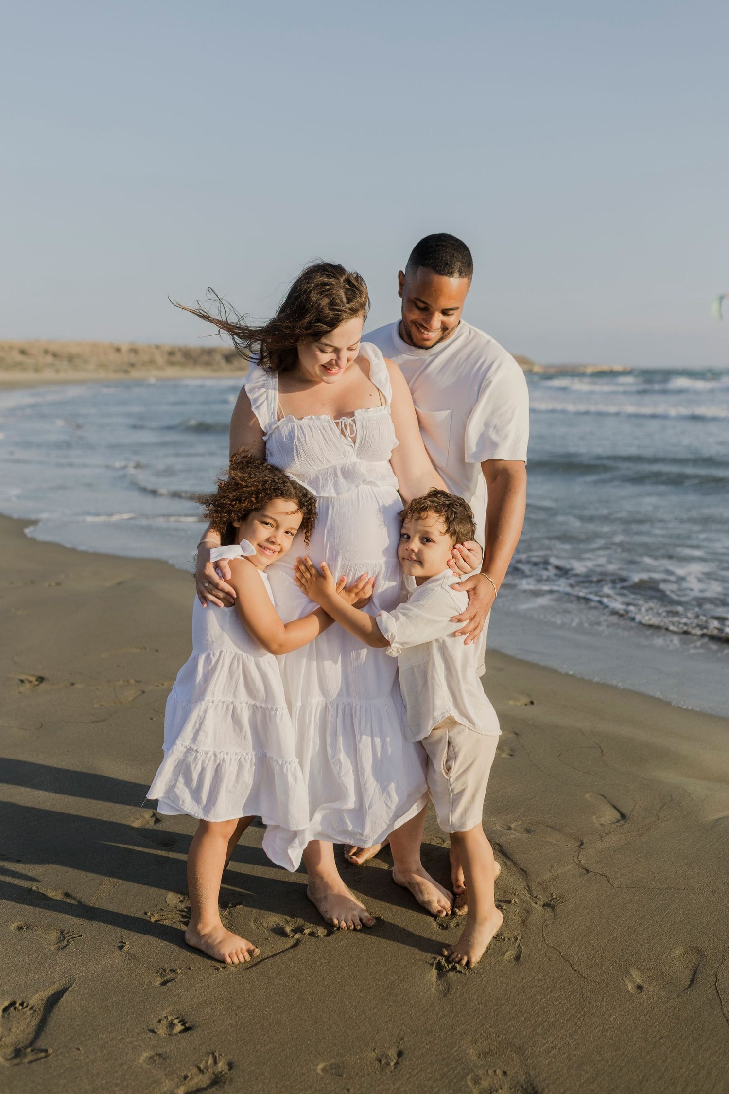 Happy family of four in white outfits posing on a sunny beach, with two children hugging their pregnant mother.