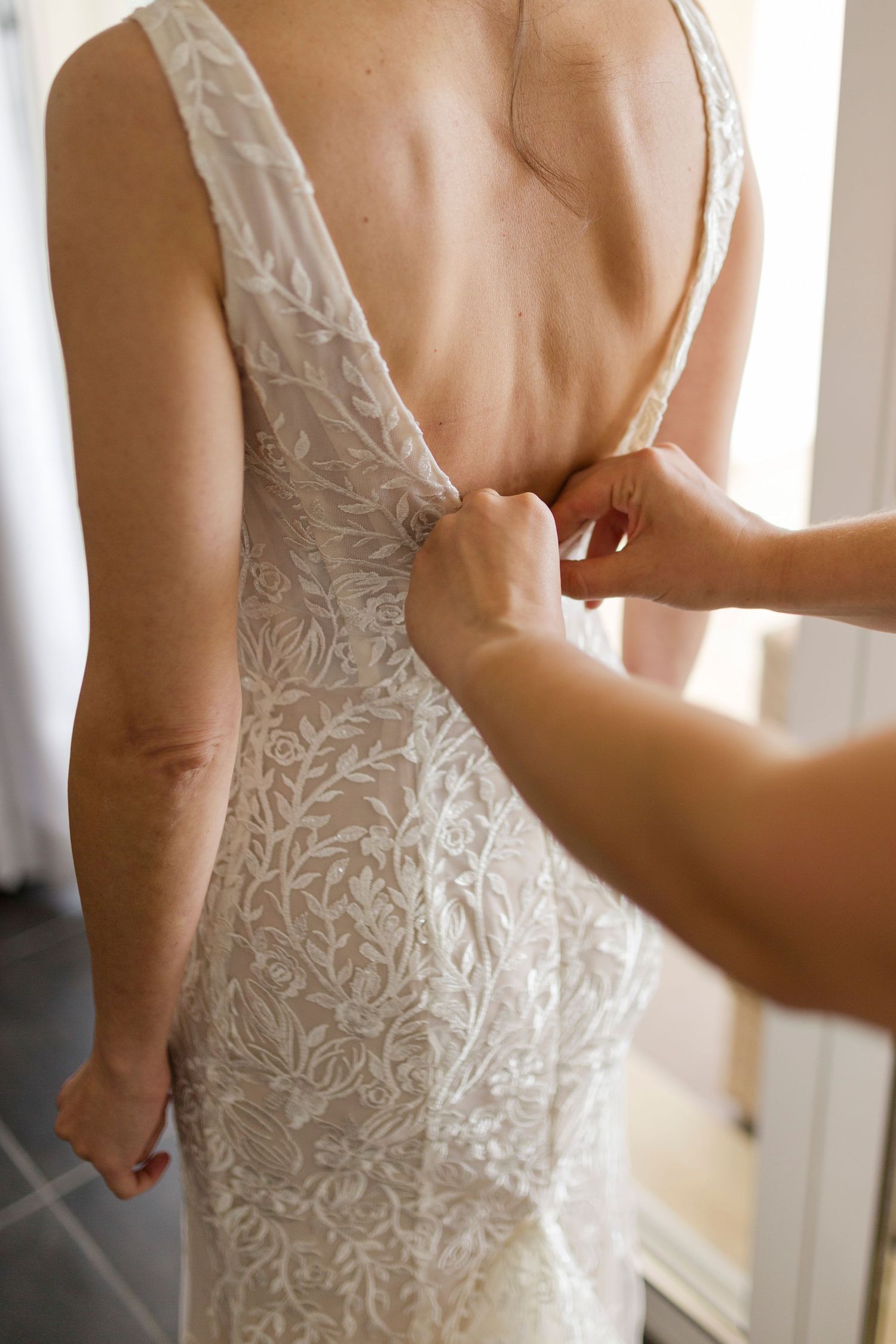 Close-up of lace wedding dress being fastened at the deep V back by bridesmaid hands.