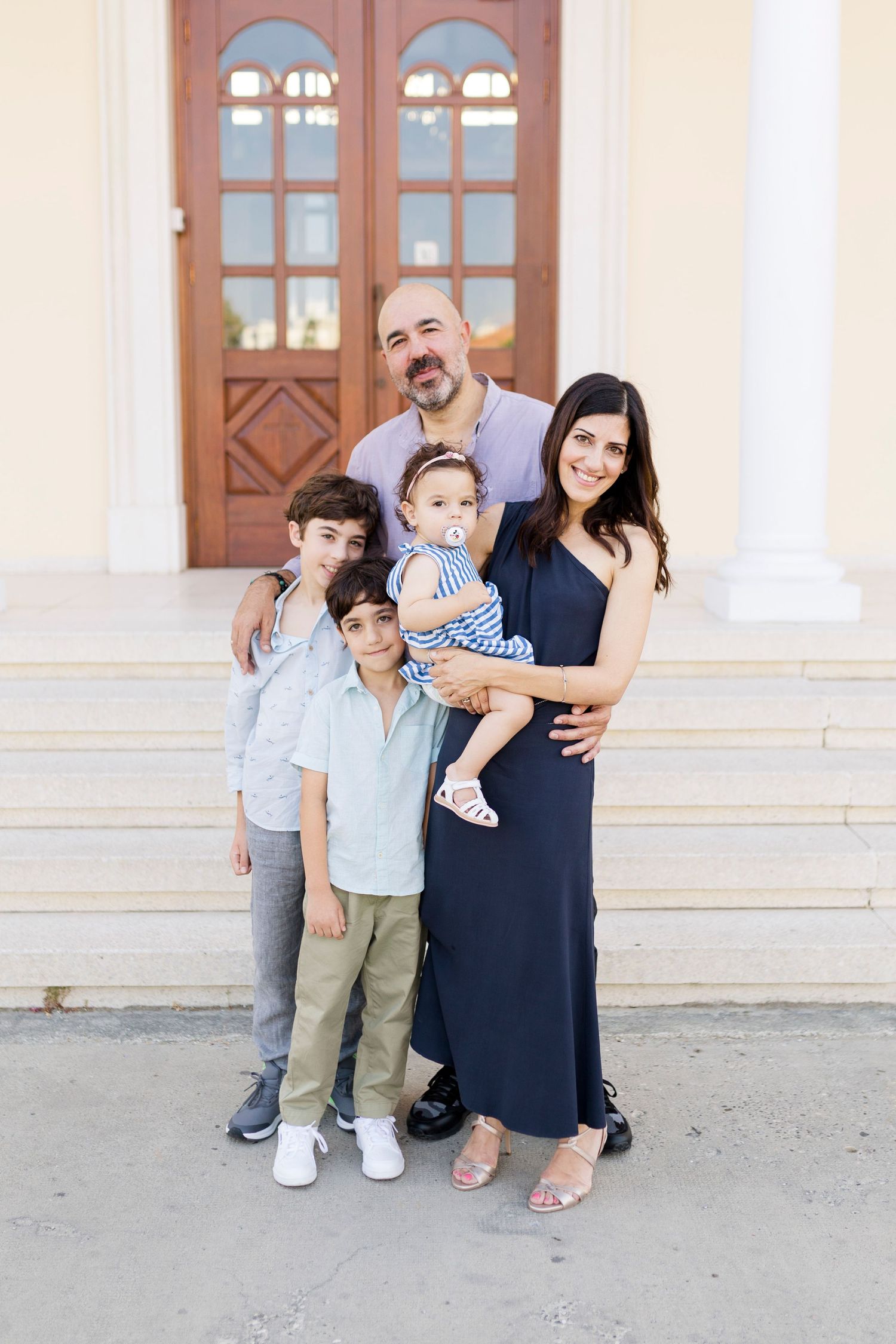 A happy family of five poses outdoors in front of a yellow building with wooden doors.
