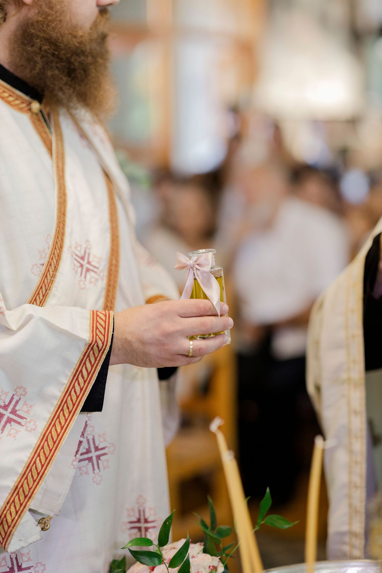 Orthodox priest in white vestments holding a small bottle of holy oil during a religious ceremony.