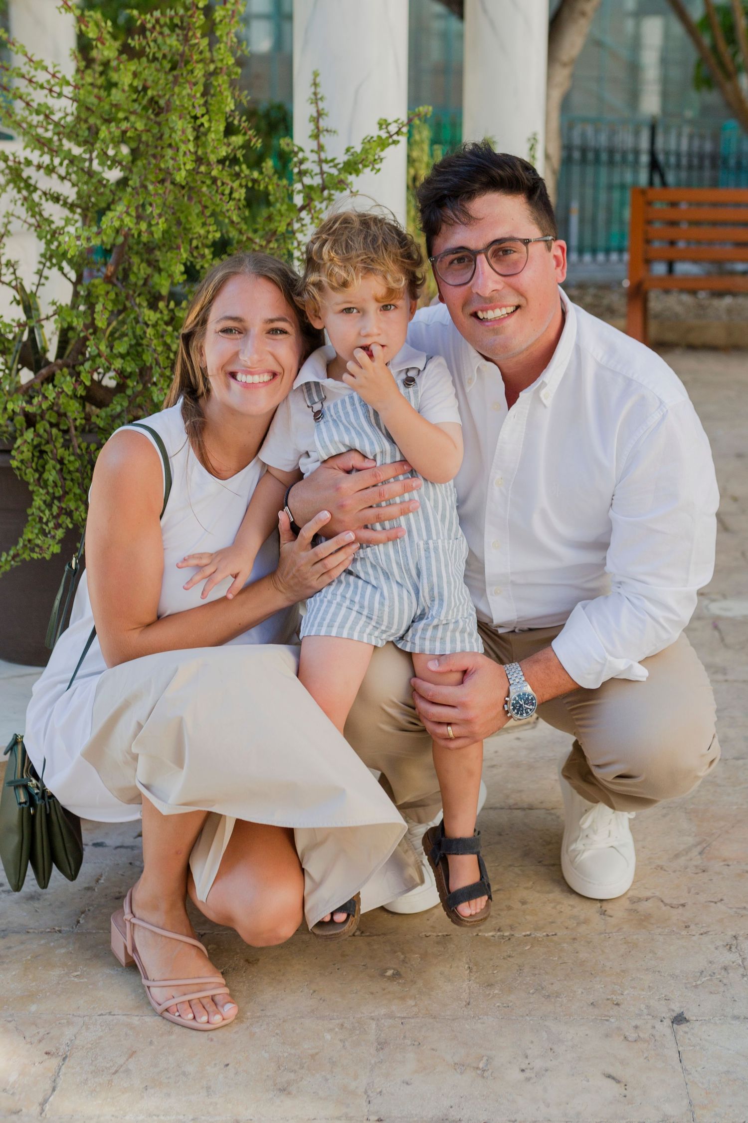 Happy family of three poses outdoors, parents crouching beside their young toddler son.
