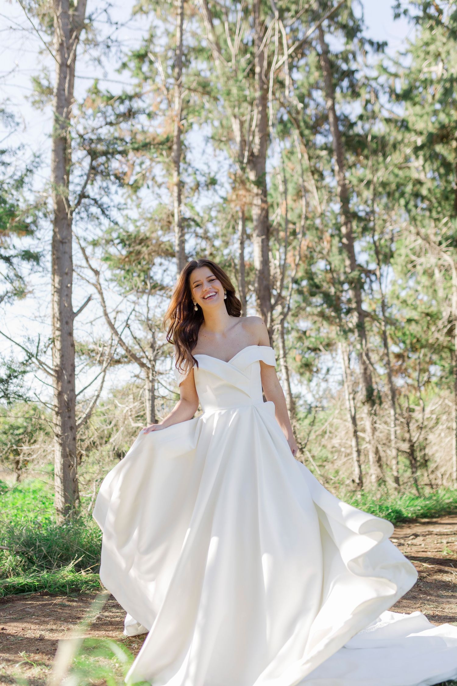 Smiling bride in an off-shoulder white satin ball gown twirling outdoors in a pine forest.