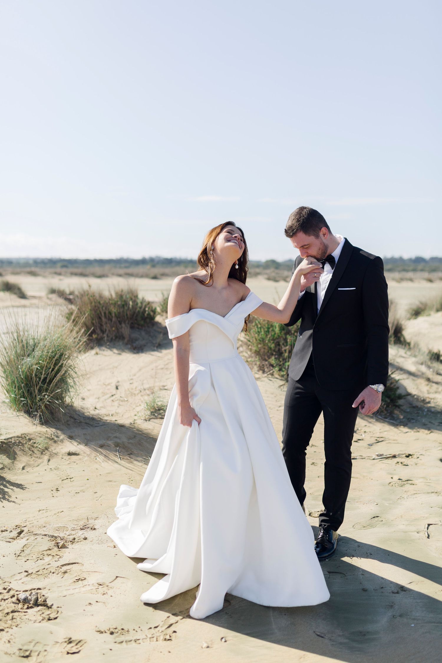 Bride in off-shoulder white gown laughs as groom in black tuxedo kisses her hand on sandy beach.