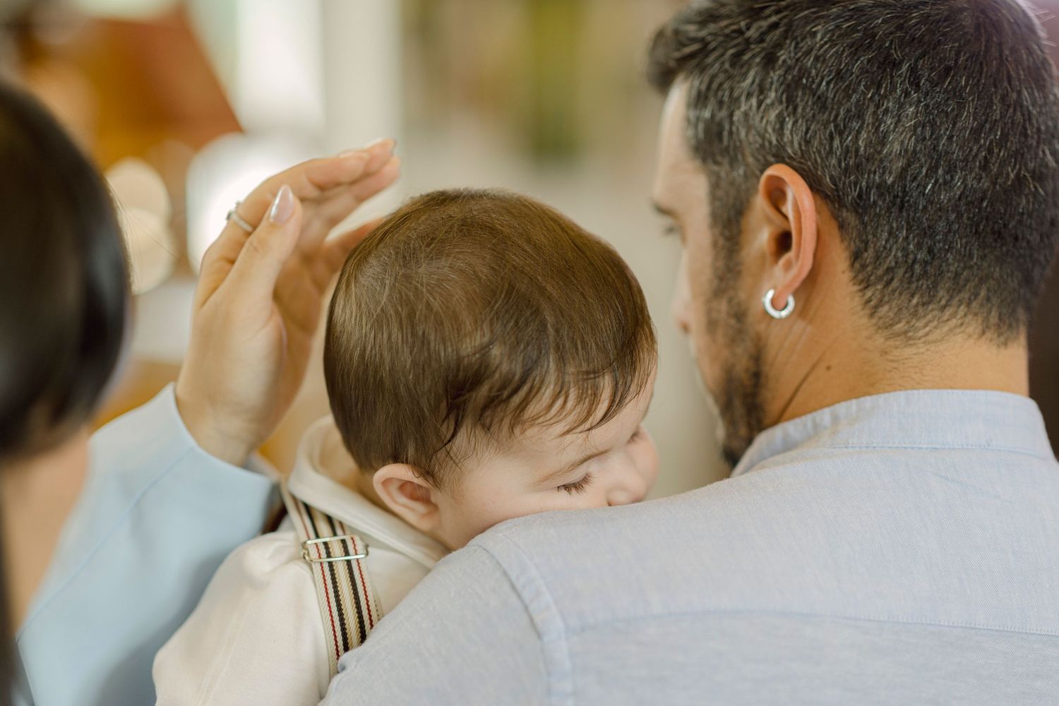 A bearded father with an earring tenderly holds a sleeping baby against his shoulder while a woman gently touches the infant's head.