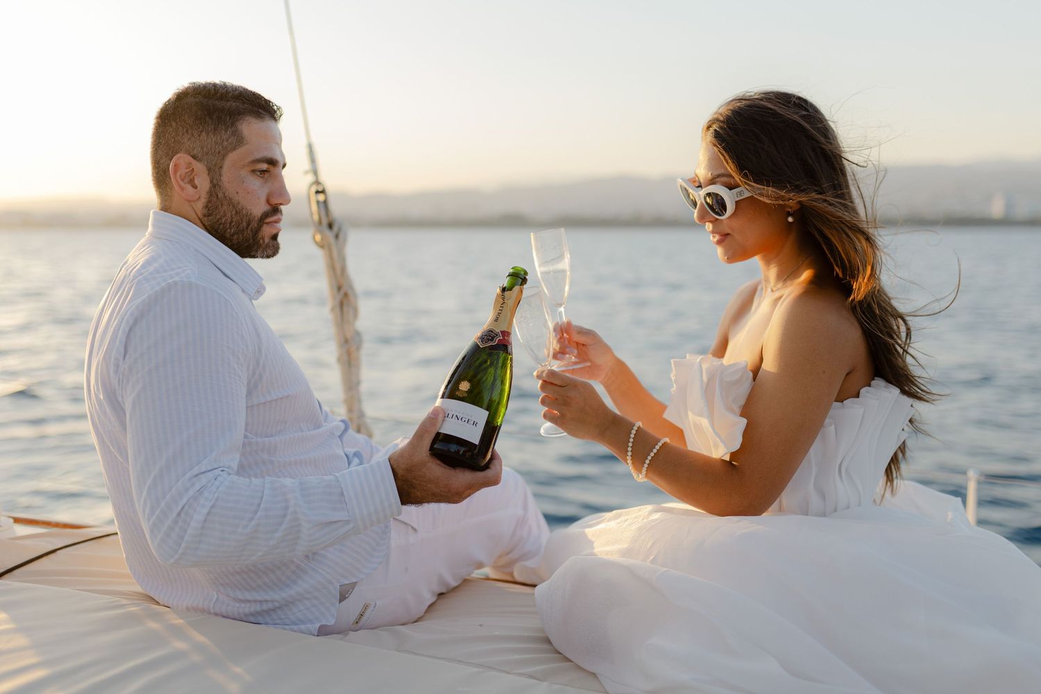 Couple in white outfits toasting with champagne on a sailboat at sunset over calm blue water.
