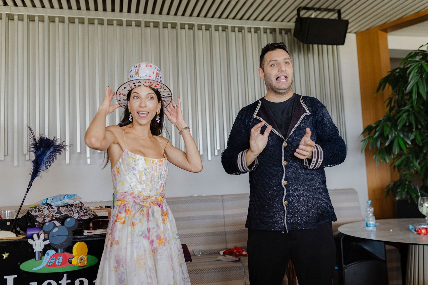 A woman in a floral dress wearing a decorative hat stands beside a smiling man in a dark cardigan at an indoor event.