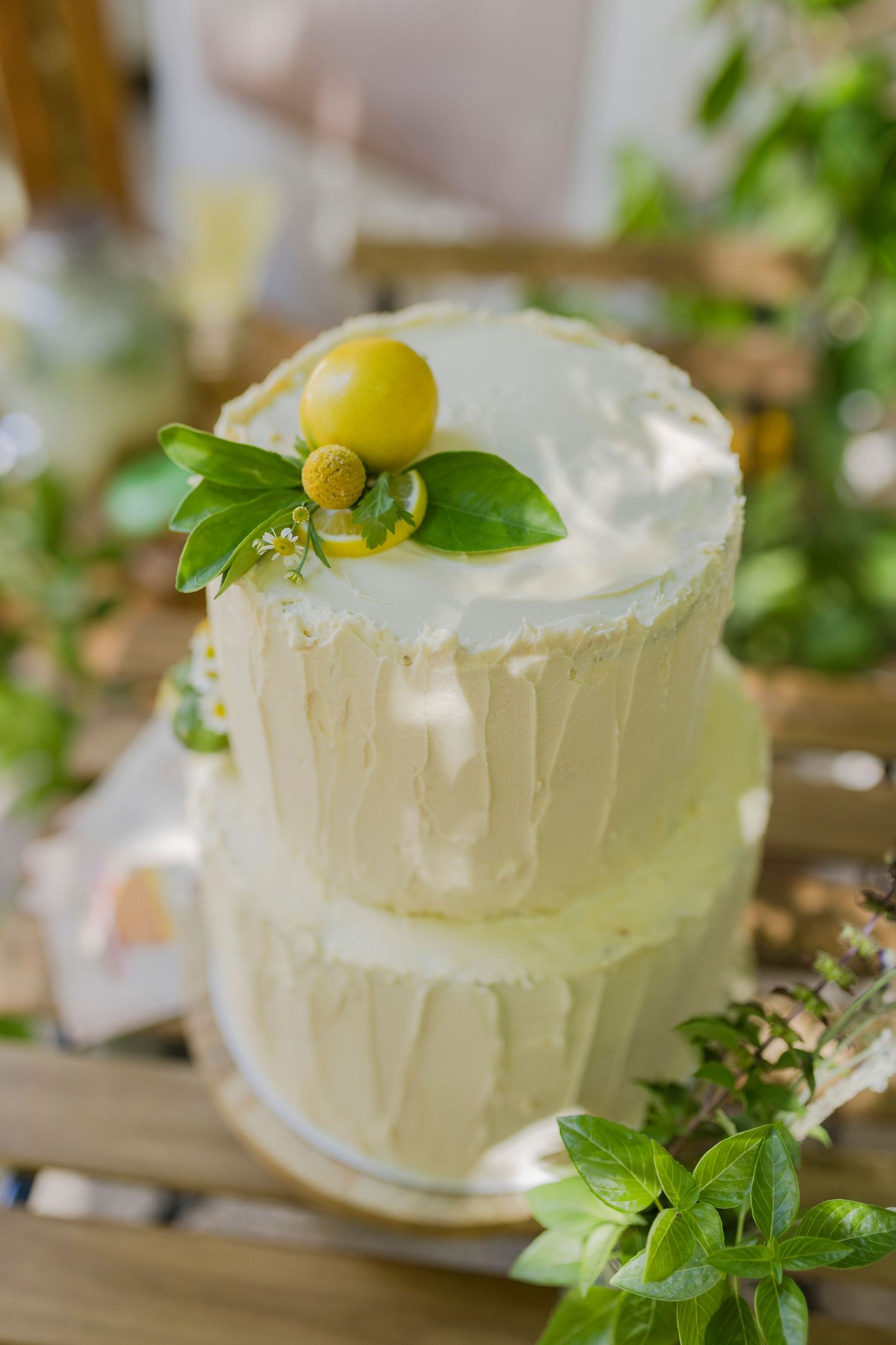 Two-tier white frosted wedding cake topped with yellow berries and fresh green leaves, surrounded by lush greenery.