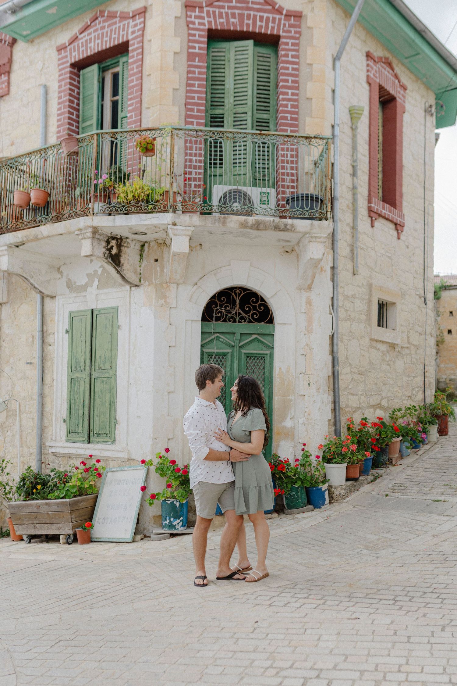 Couple holding hands in front of a charming stone building with green shutters and flower pots.