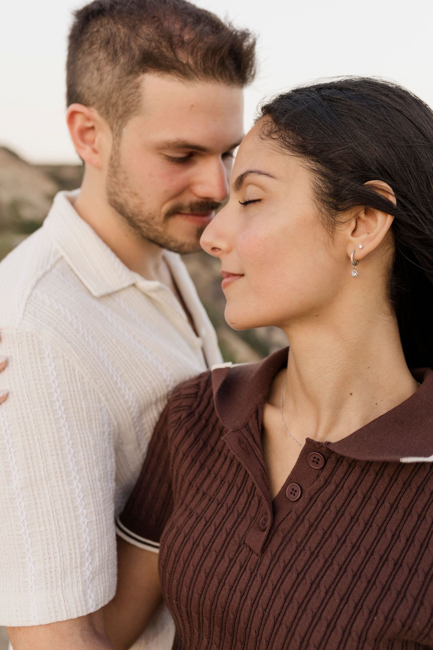 A couple shares a tender moment with eyes closed, the woman in brown and man in white shirt embracing outdoors.
