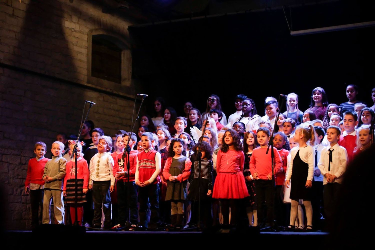 Children and adults perform on stage in red and black outfits during a school choir concert under dramatic lighting.