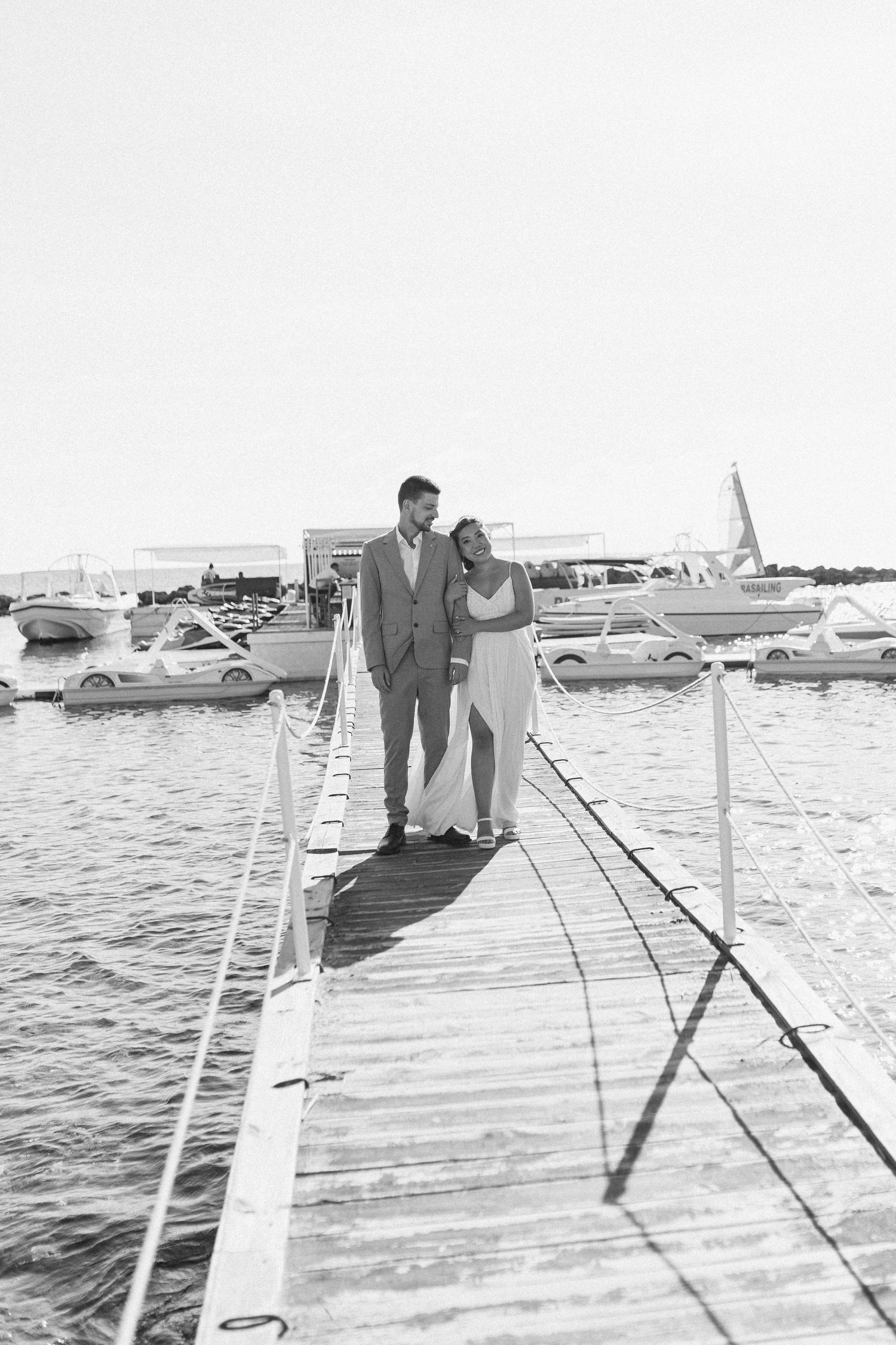 Black and white photo of bride and groom embracing on a wooden dock surrounded by boats.
