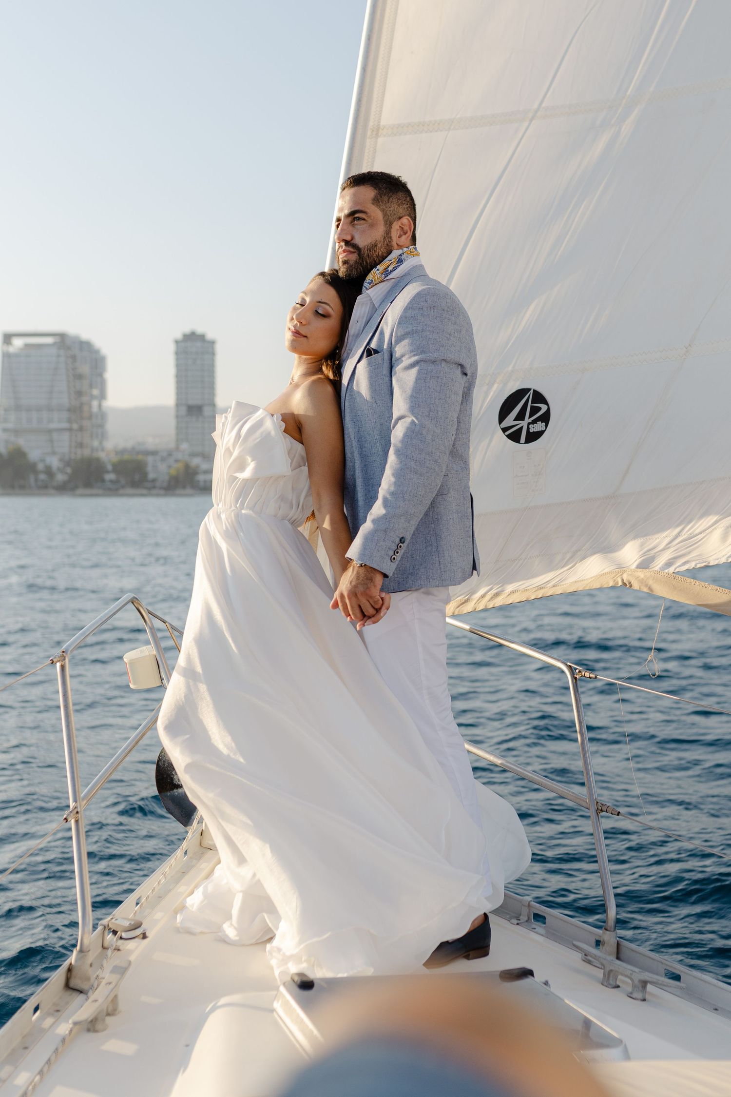 Elegant couple in wedding attire embracing on a white sailboat with ocean and city skyline in background.