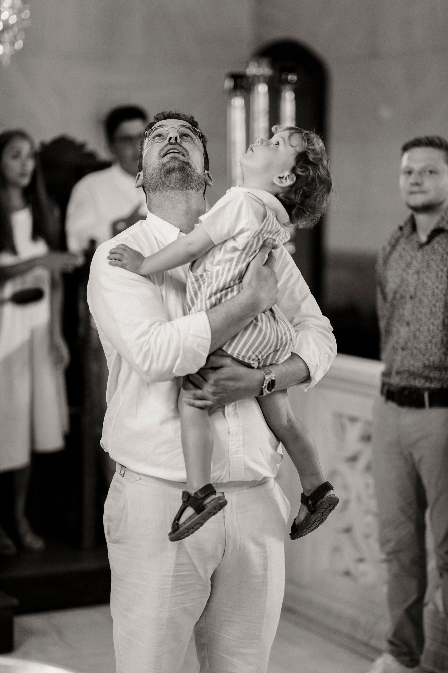 Black and white photo of a bearded man lifting a young child in a church setting during a ceremony.