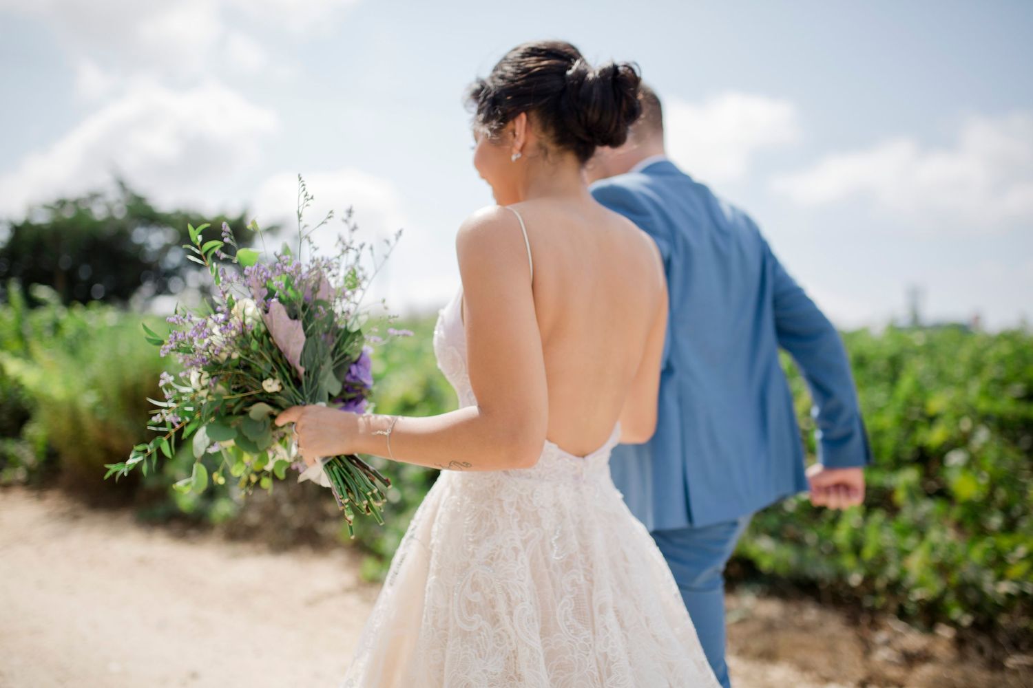 Bride in backless lace gown holding purple wildflower bouquet walks with groom in blue suit at outdoor vineyard wedding.