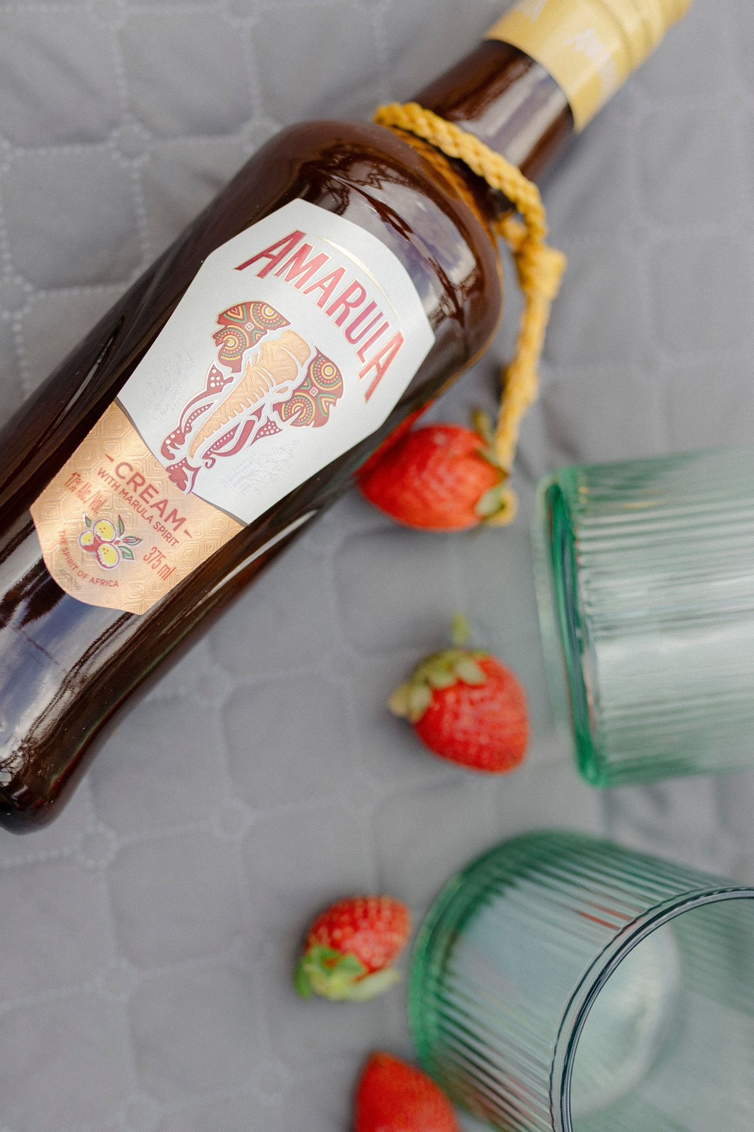 Amarula cream liqueur bottle lying on gray surface with fresh strawberries and two green ribbed glasses nearby.