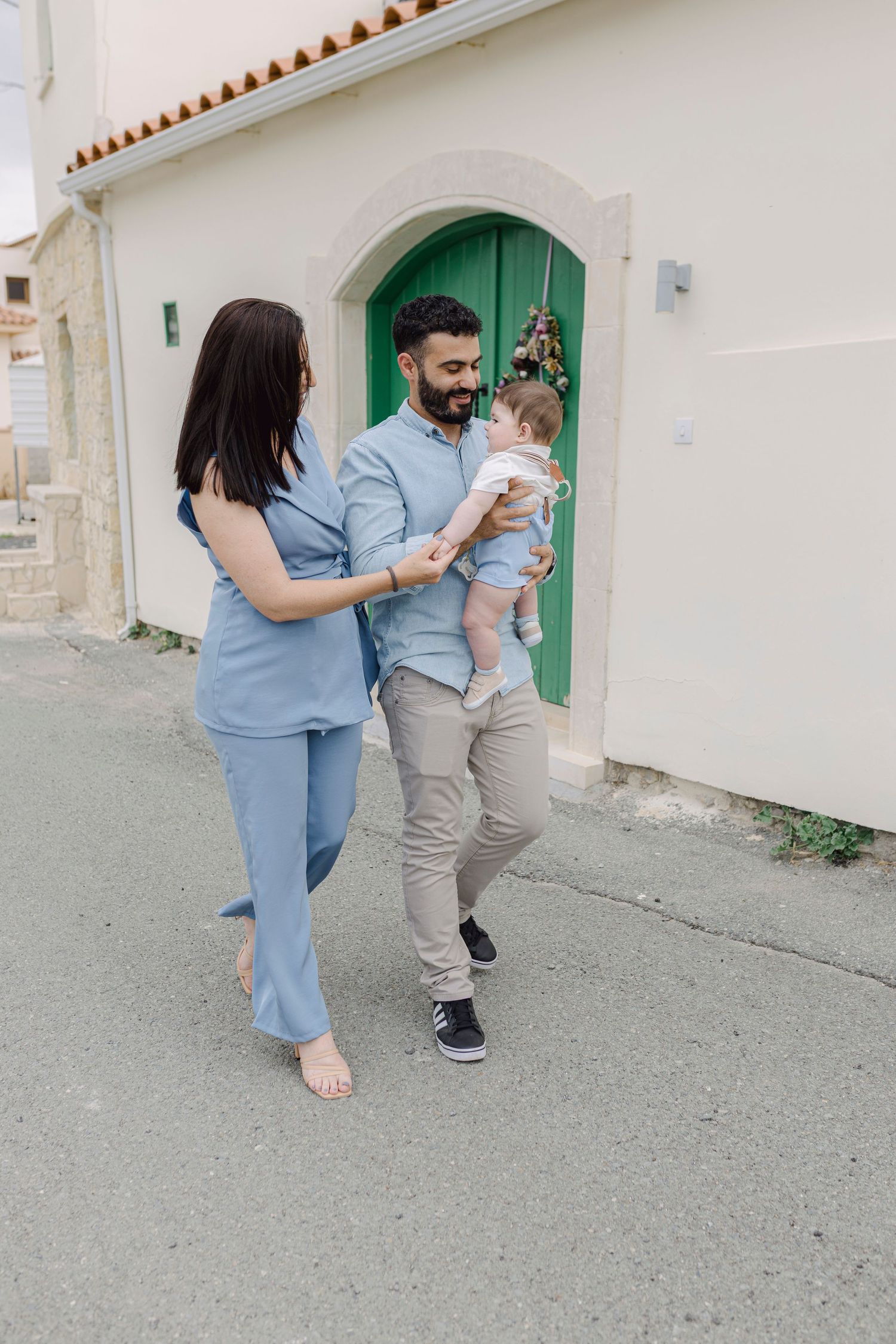 Young family with baby walking outside white building with green arched door, parents in blue outfits.
