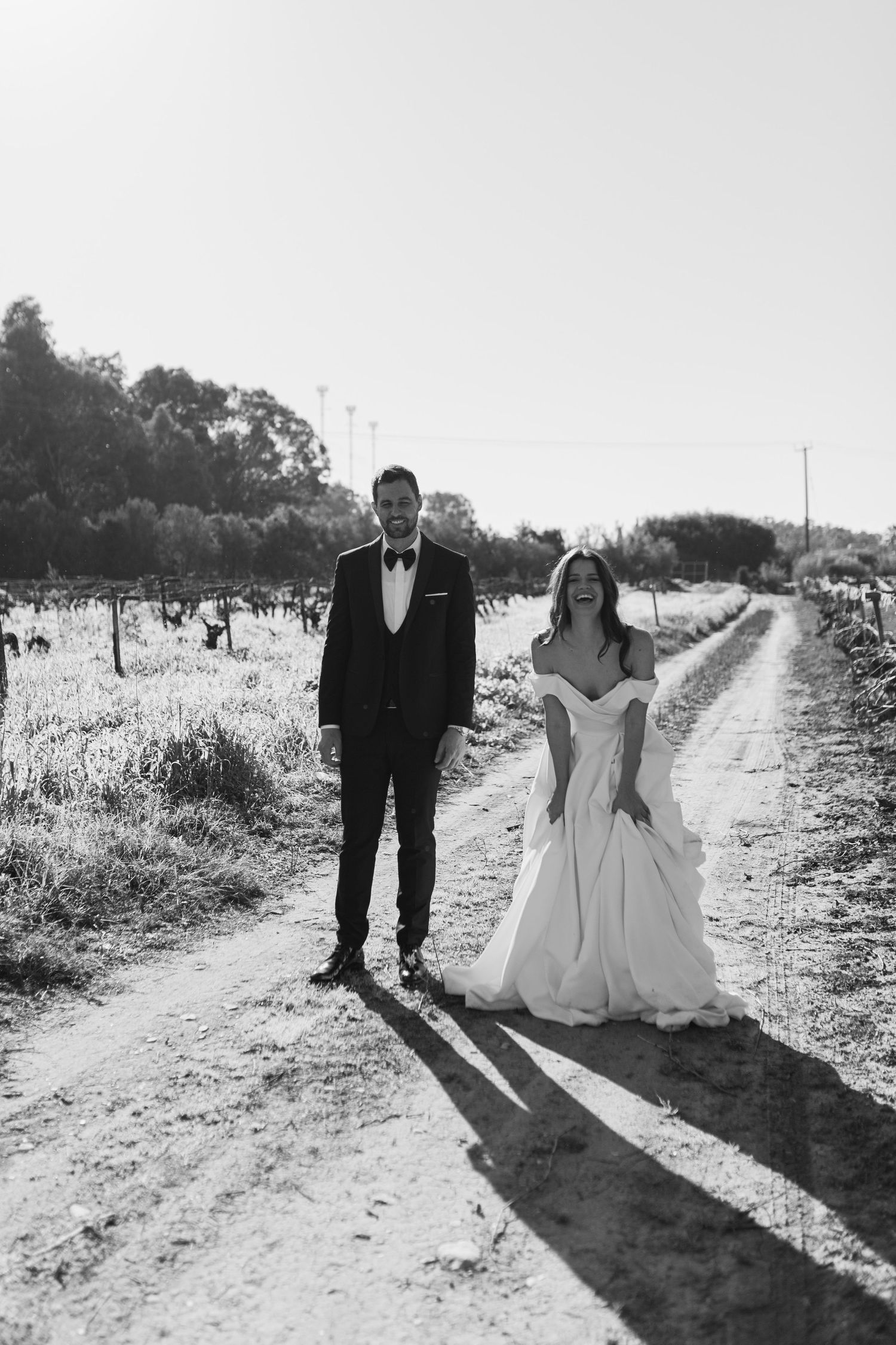 Black and white photo of bride and groom walking hand in hand down a rural dirt road at sunset.
