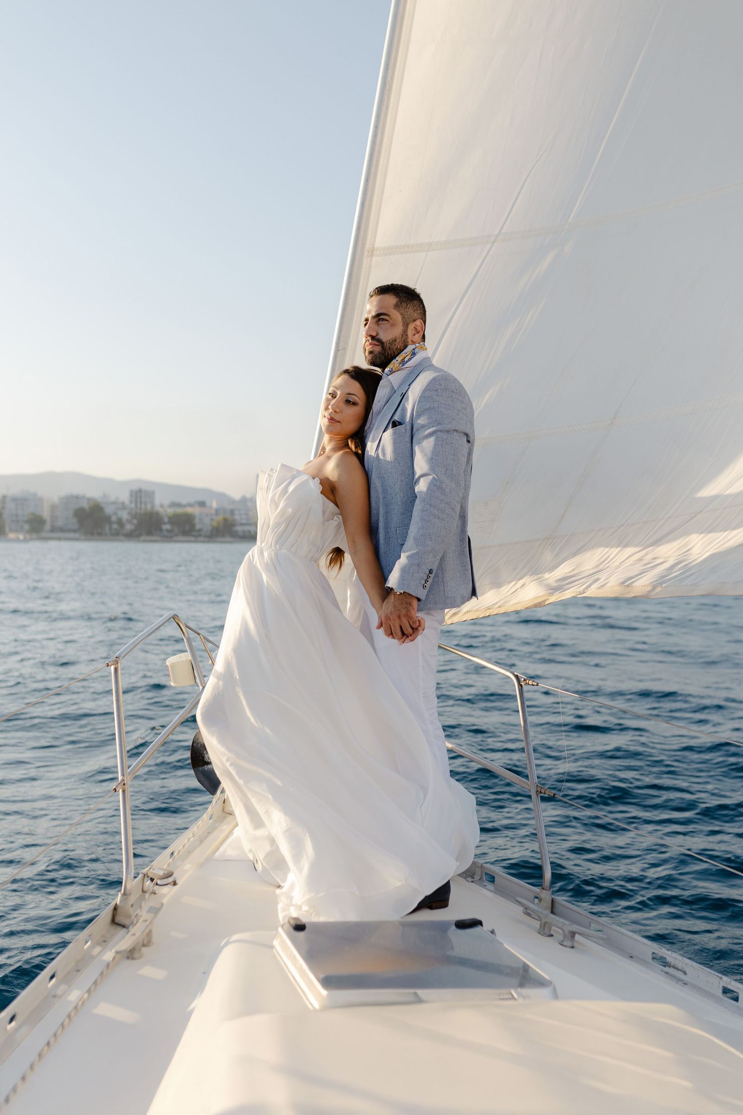 Bride and groom embracing on the bow of a white sailboat on calm blue ocean waters.