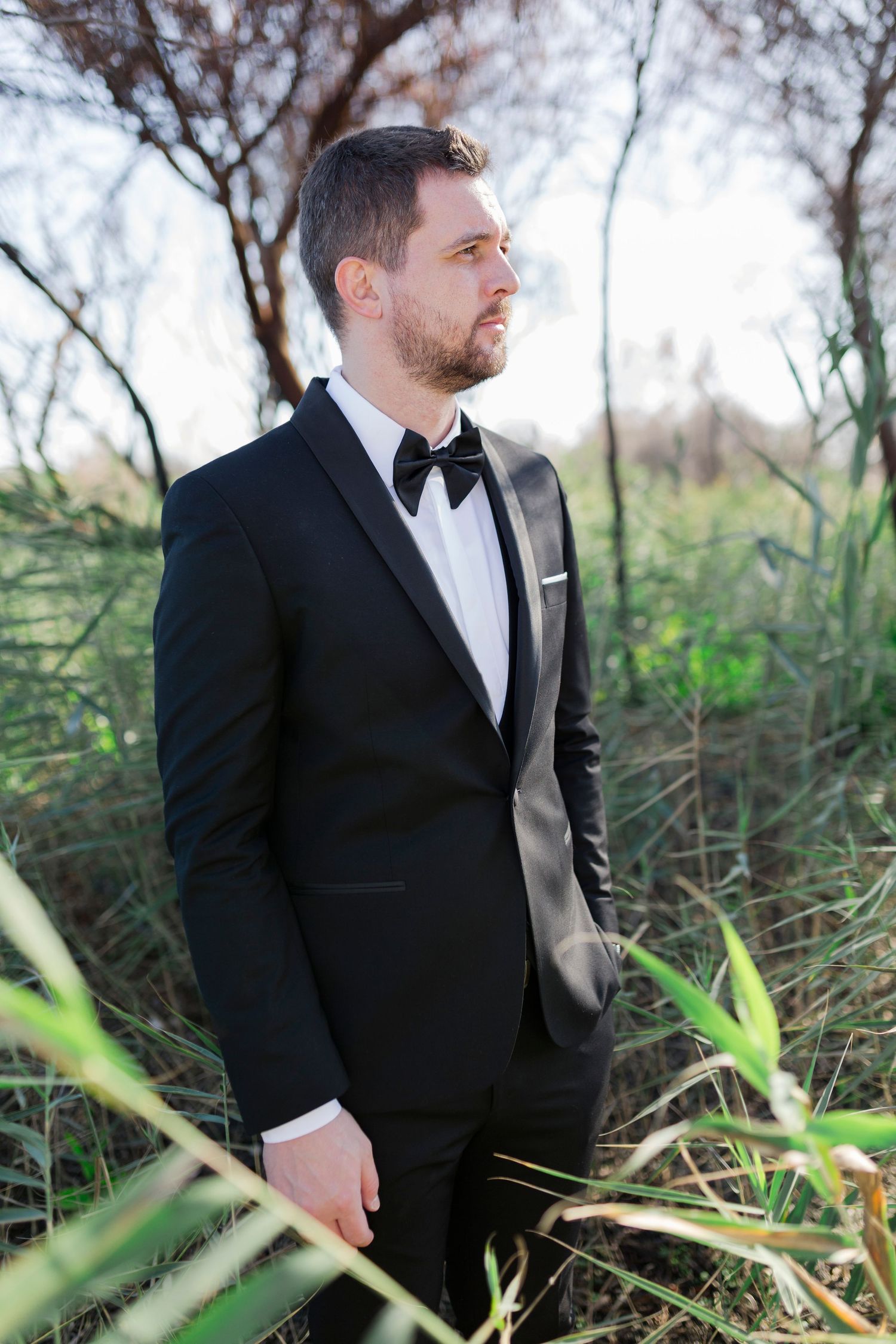 Elegant groom in black tuxedo with bow tie standing outdoors among tall green grass and trees.