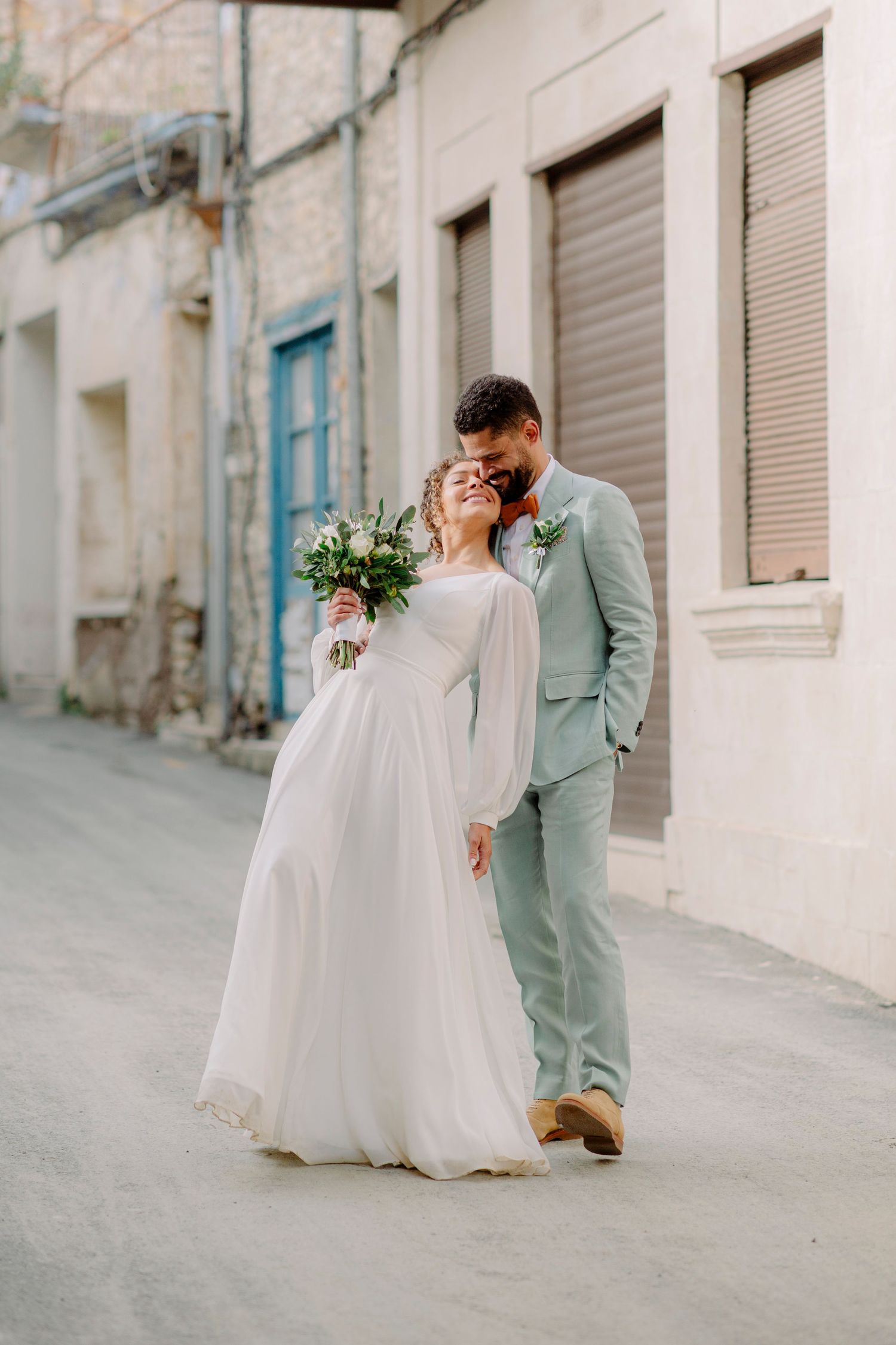 A bride and groom walk together on a charming street, she holds a bouquet in a white gown, he wears a sage suit.