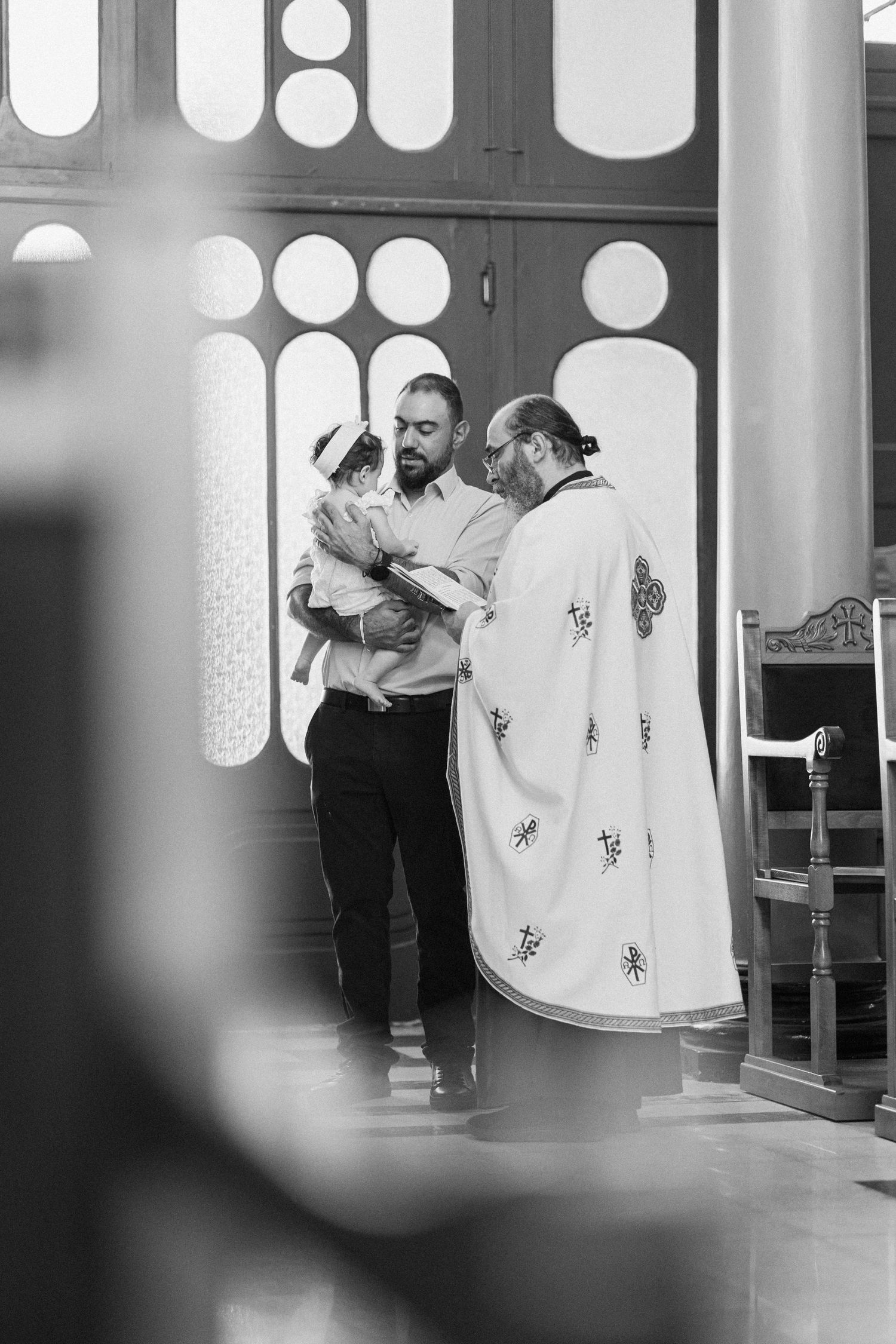 Black and white photo of a priest blessing a man holding a baby during a baptism ceremony in a church.