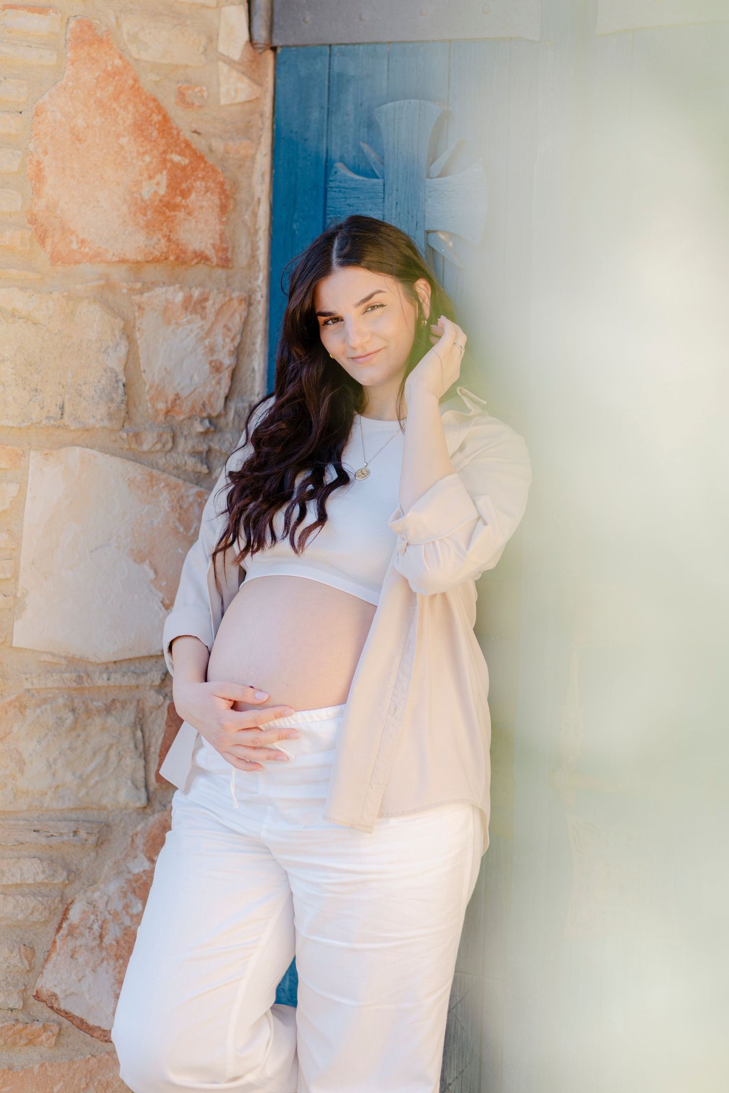 Pregnant woman in white outfit cradling her baby bump, leaning against a stone wall with a blue door.