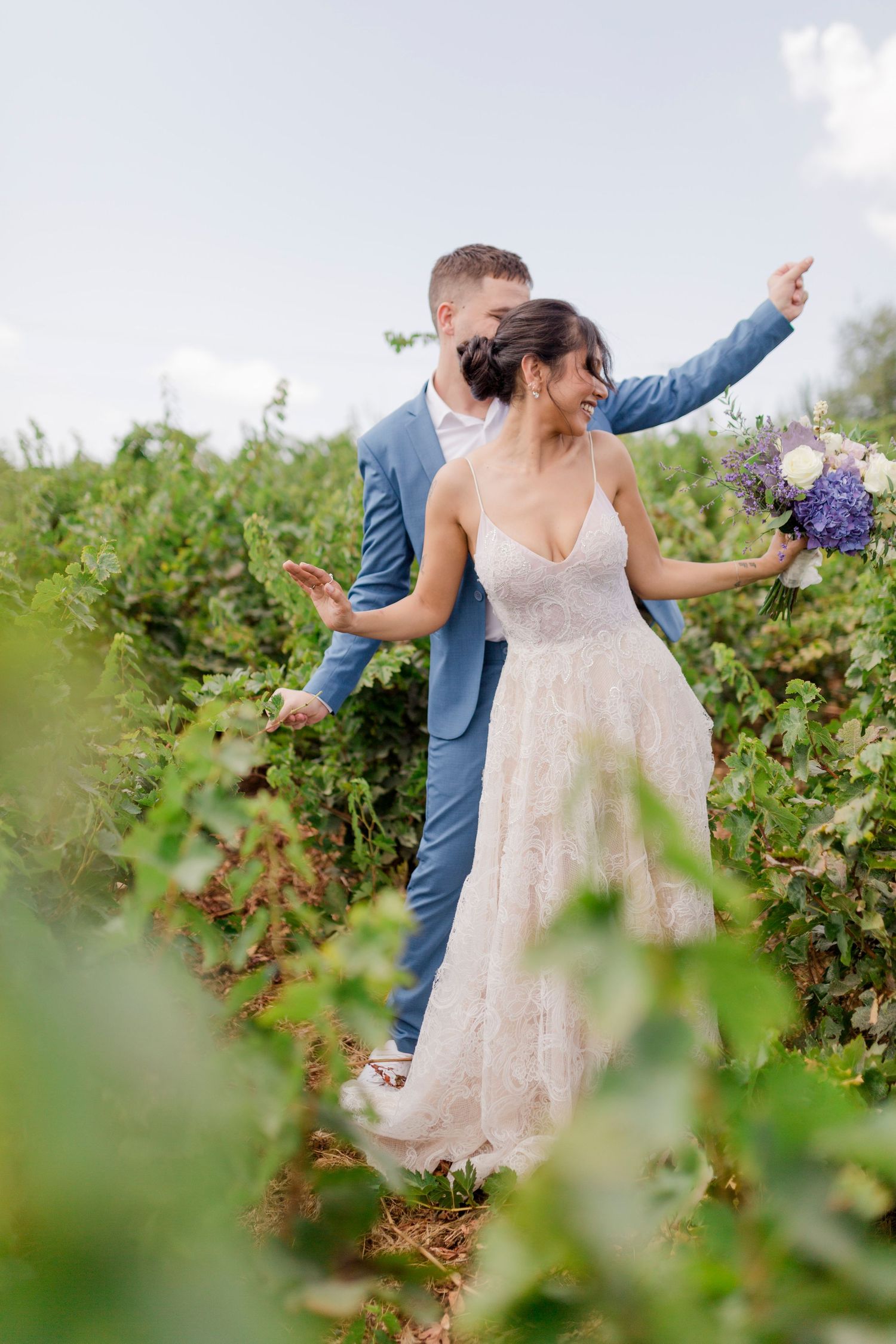 Bride and groom posing joyfully in a lush vineyard, bride holding purple and white bouquet in lace gown.