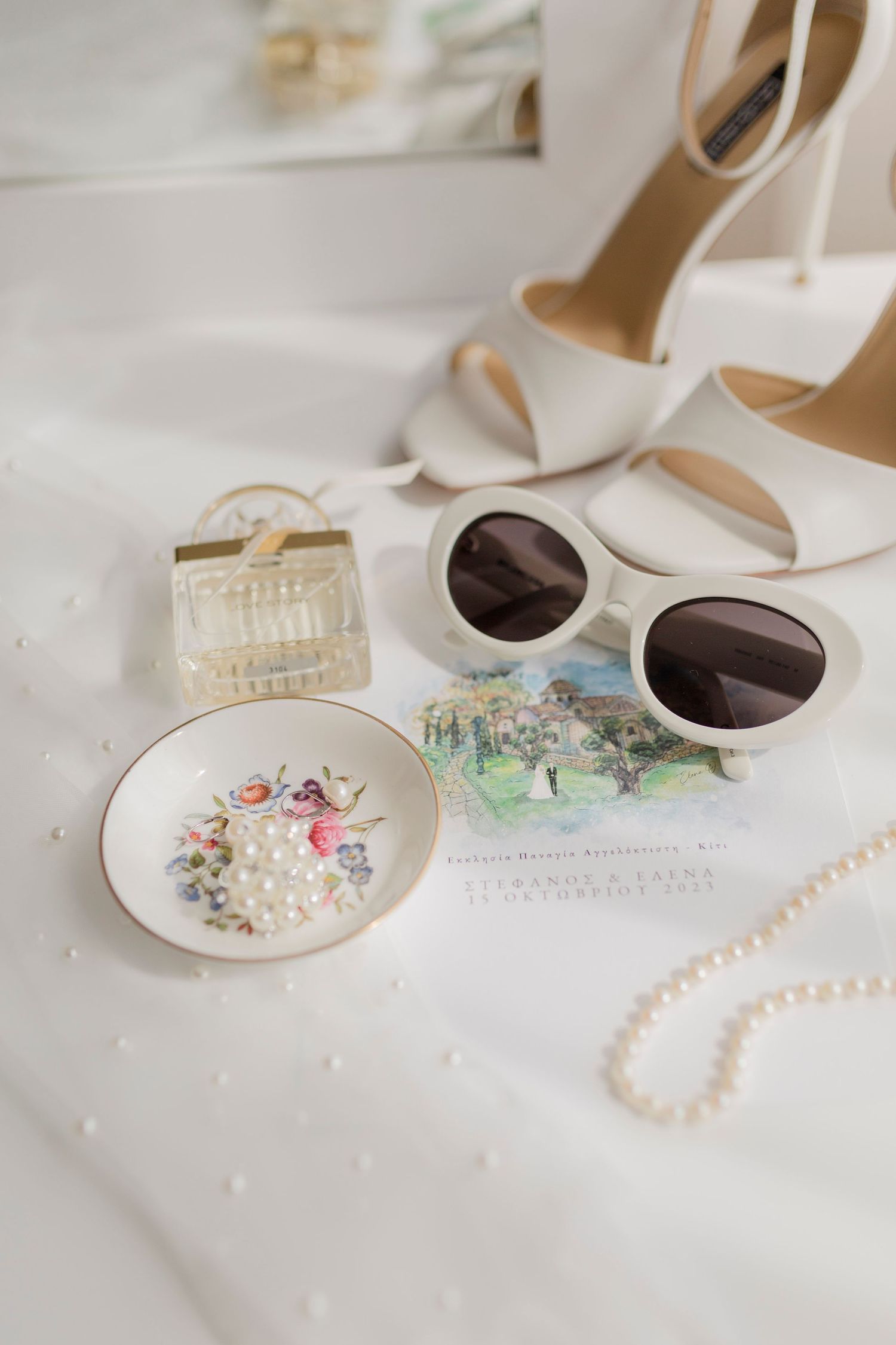Flat lay of bridal accessories including white heels, sunglasses, pearl necklace, and floral dish.