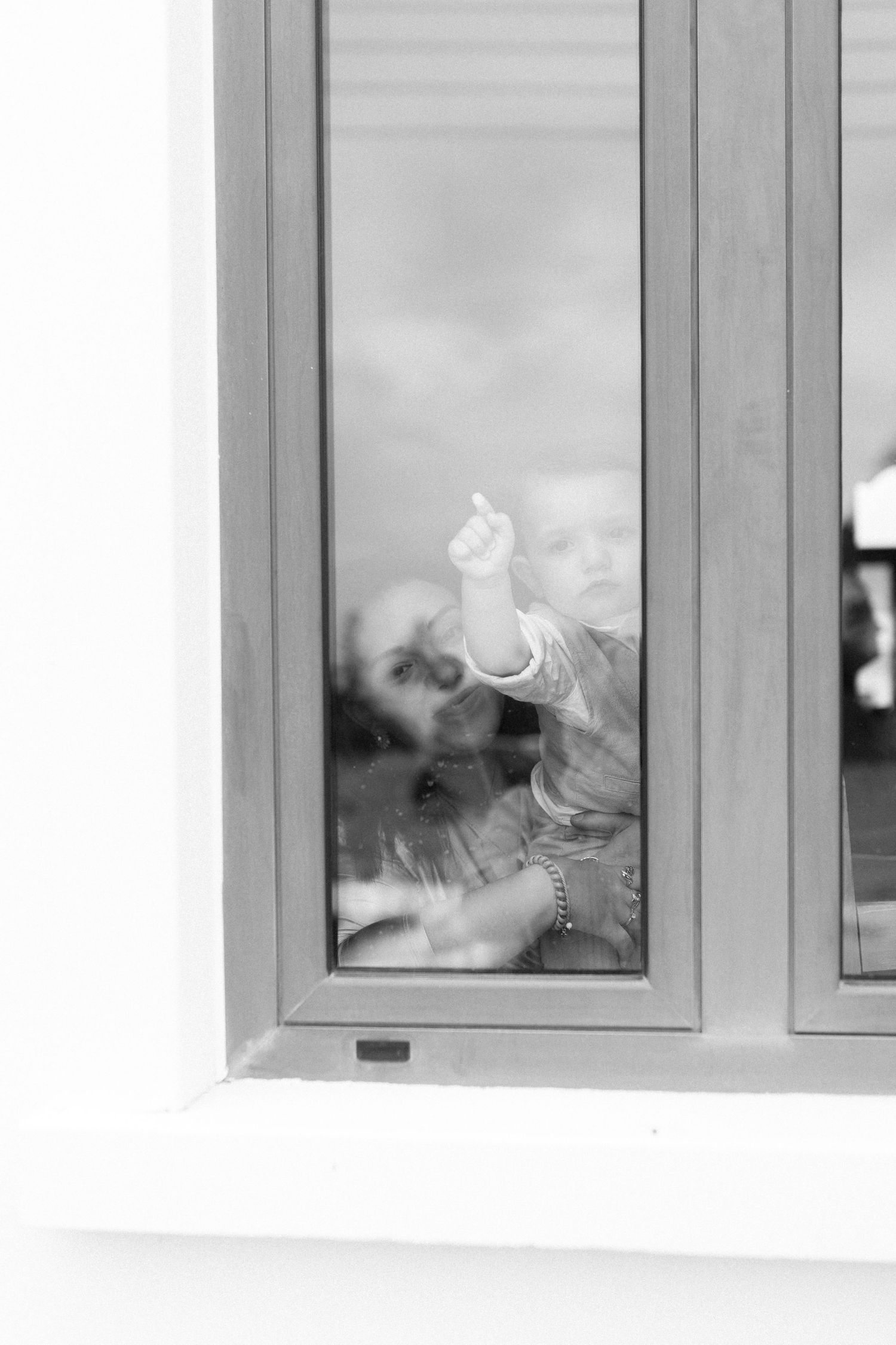 Black and white photo of adult holding baby near window, child pointing finger upward indoors.