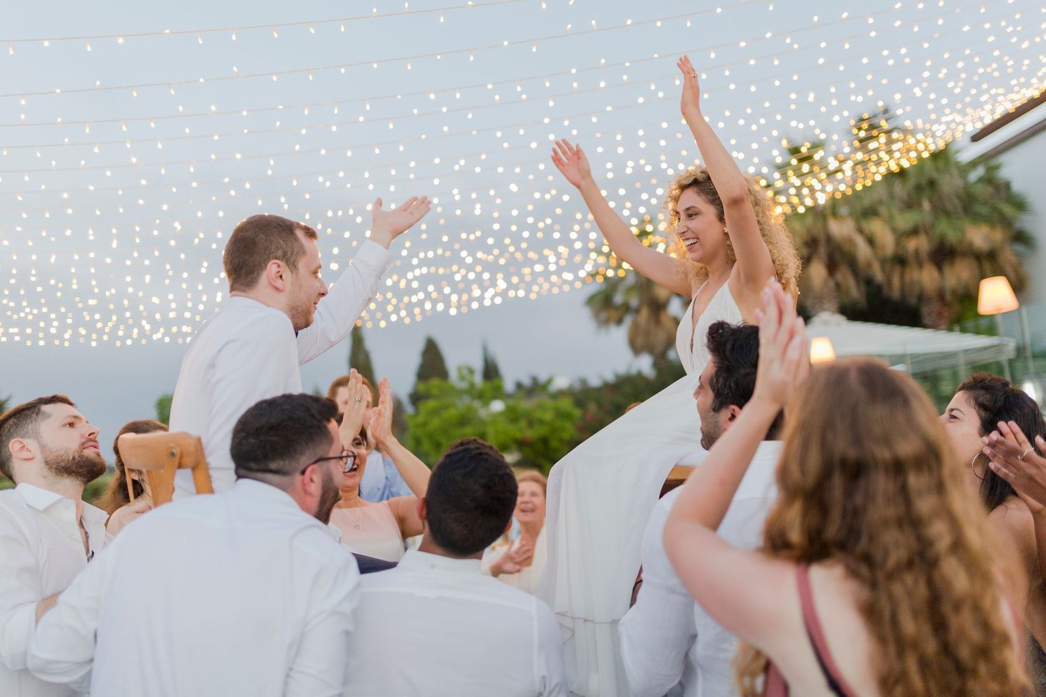 Bride and groom celebrate joyfully at outdoor wedding reception with string lights and cheering guests.