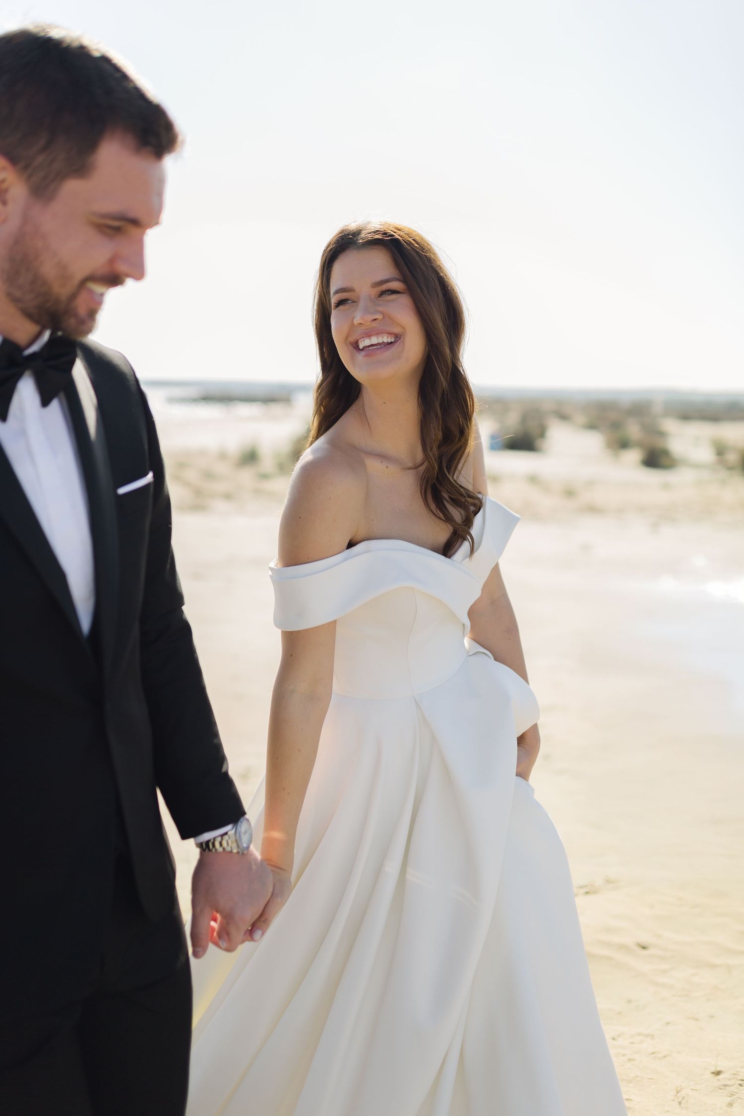 Smiling bride in off-shoulder white gown holds hands with groom in black tuxedo on sandy beach.