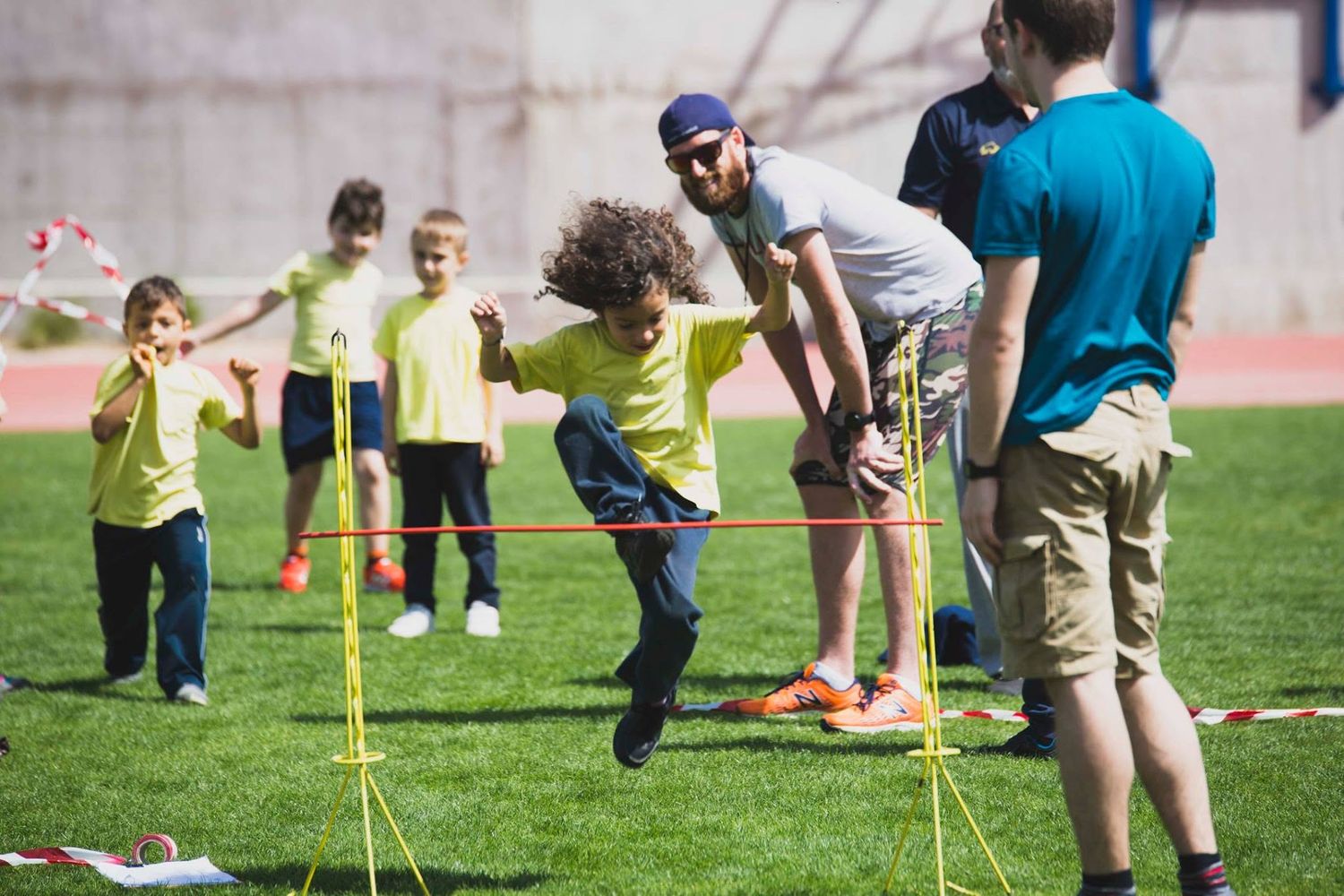 Children in yellow shirts jump rope outdoors on a green field during a fun sports day activity with adult supervisors.