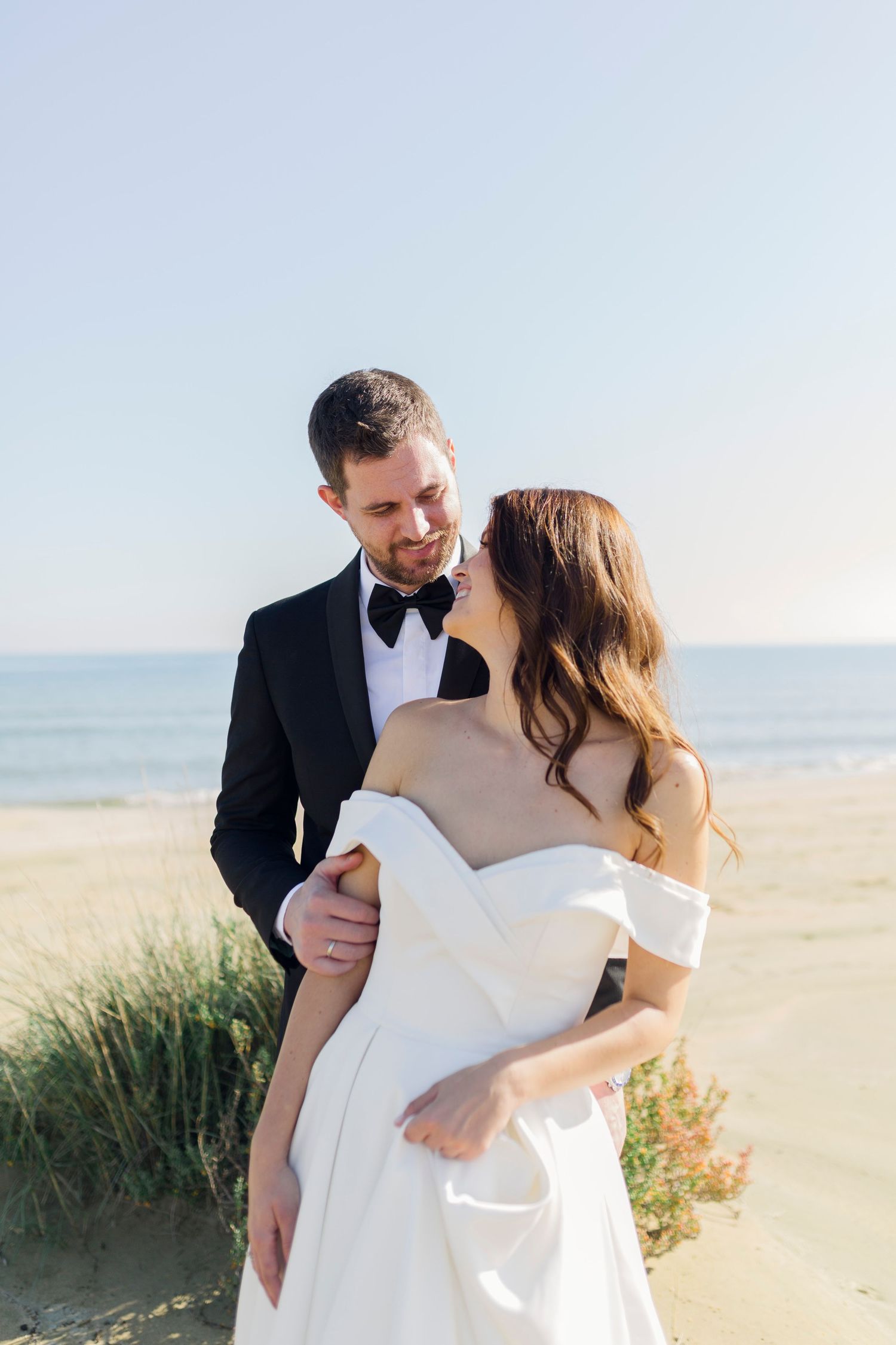 A bride and groom share a romantic moment on a sandy beach, with the ocean in the background on their wedding day.