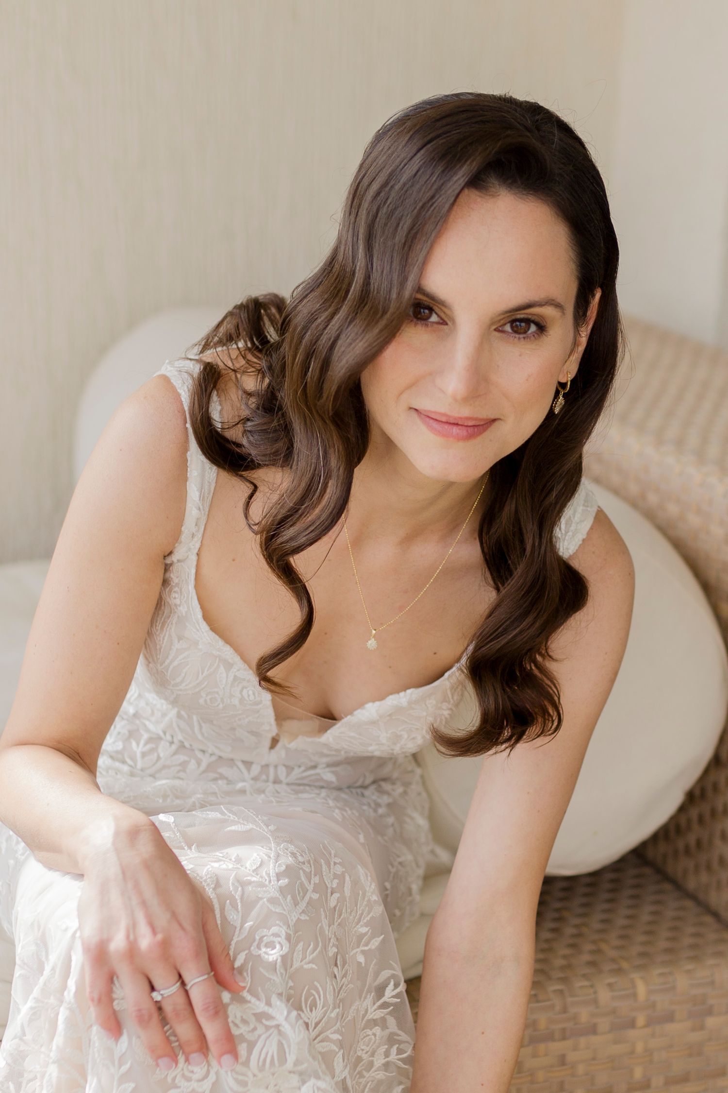 Beautiful brunette bride in a lace wedding gown sitting and smiling softly indoors.