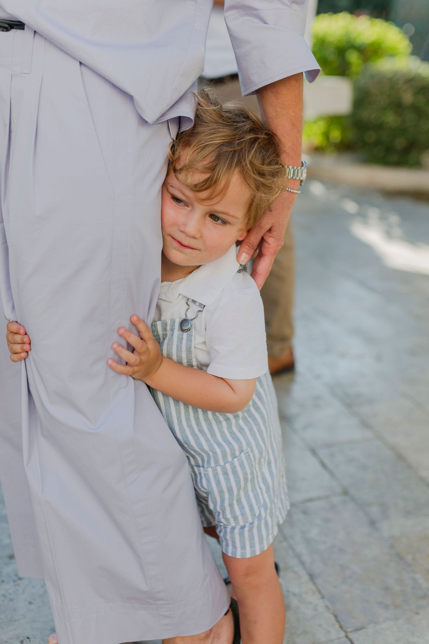 A young toddler boy hugging an adult's leg outdoors, wearing striped shorts and a white shirt.